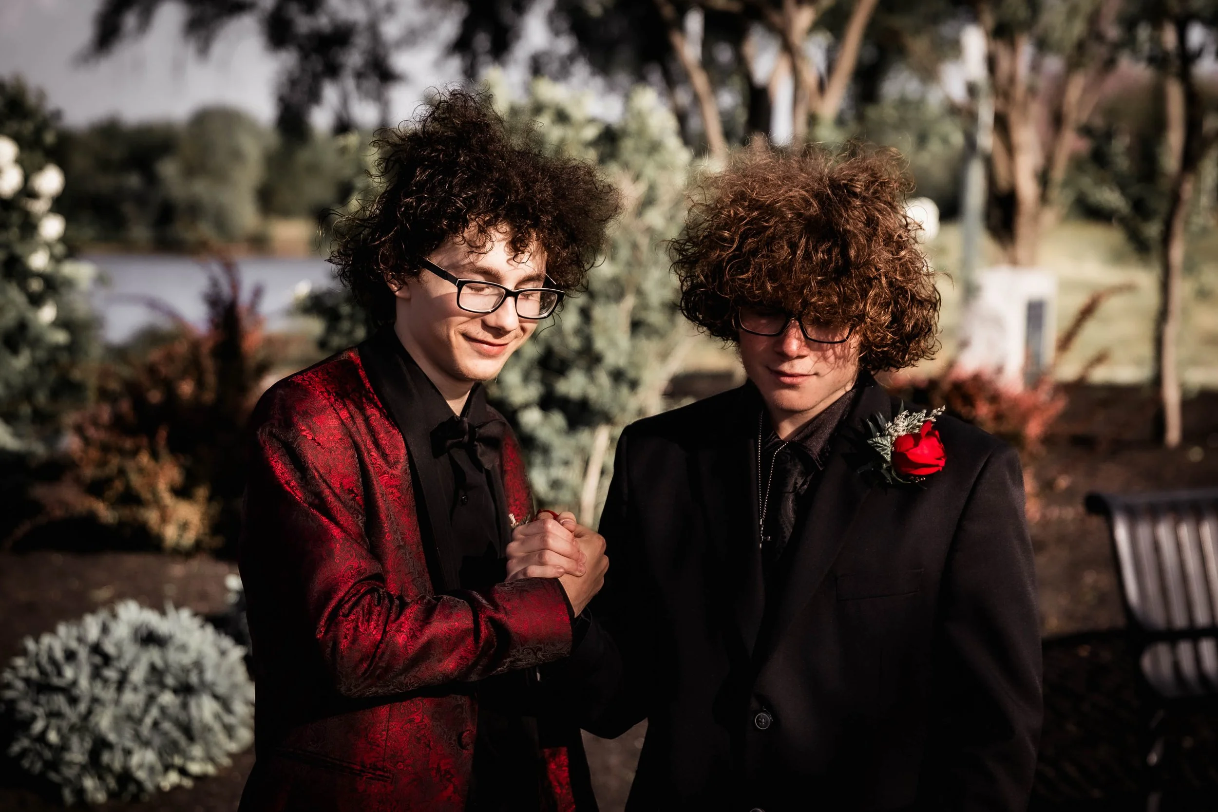 Two young men with curly hair, wearing glasses and formal jackets, holding hands and praying together outdoors during evening, with trees and bushes in the background.