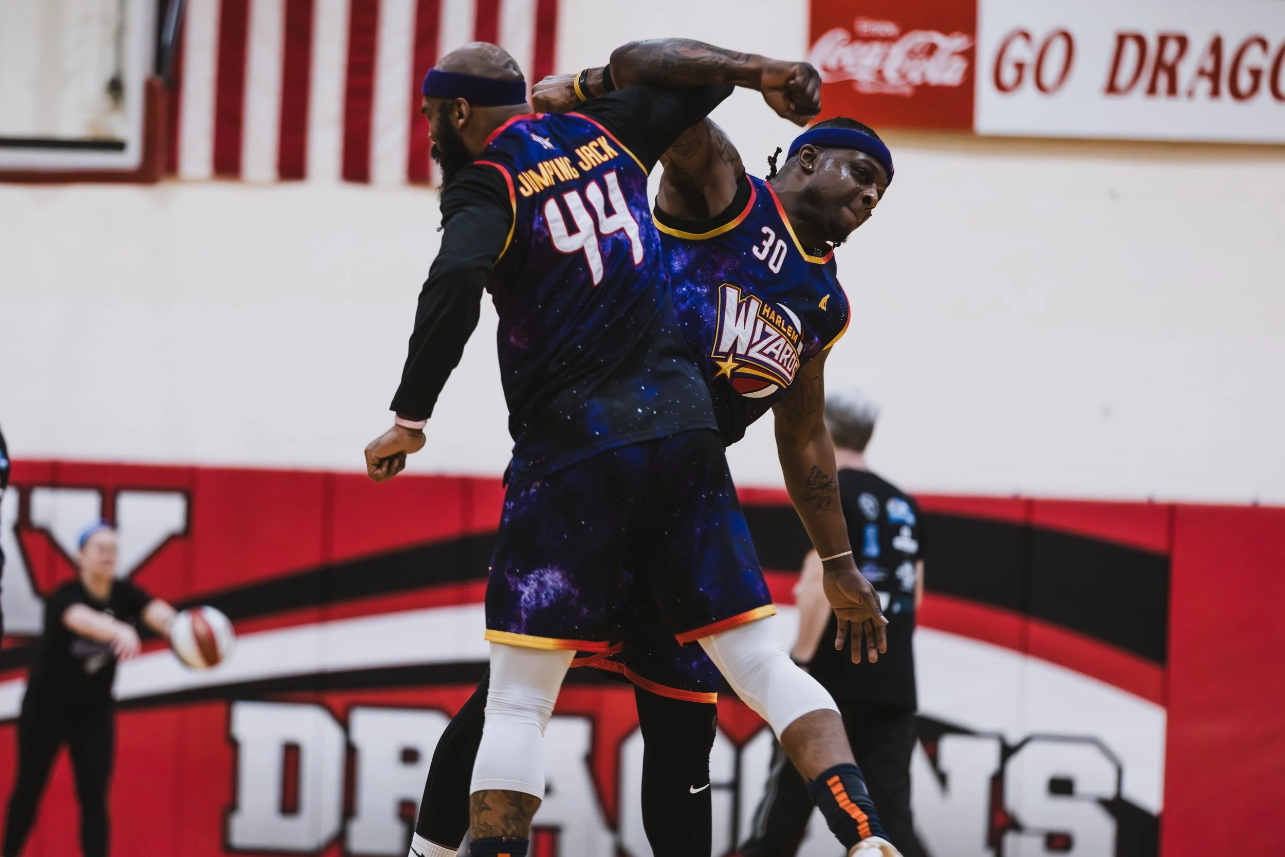 Two basketball players from the Harlem Wizards team practicing in a gym, with one player wearing a black and galaxy-patterned uniform and the other in similar attire, and a coach or trainer in the background.