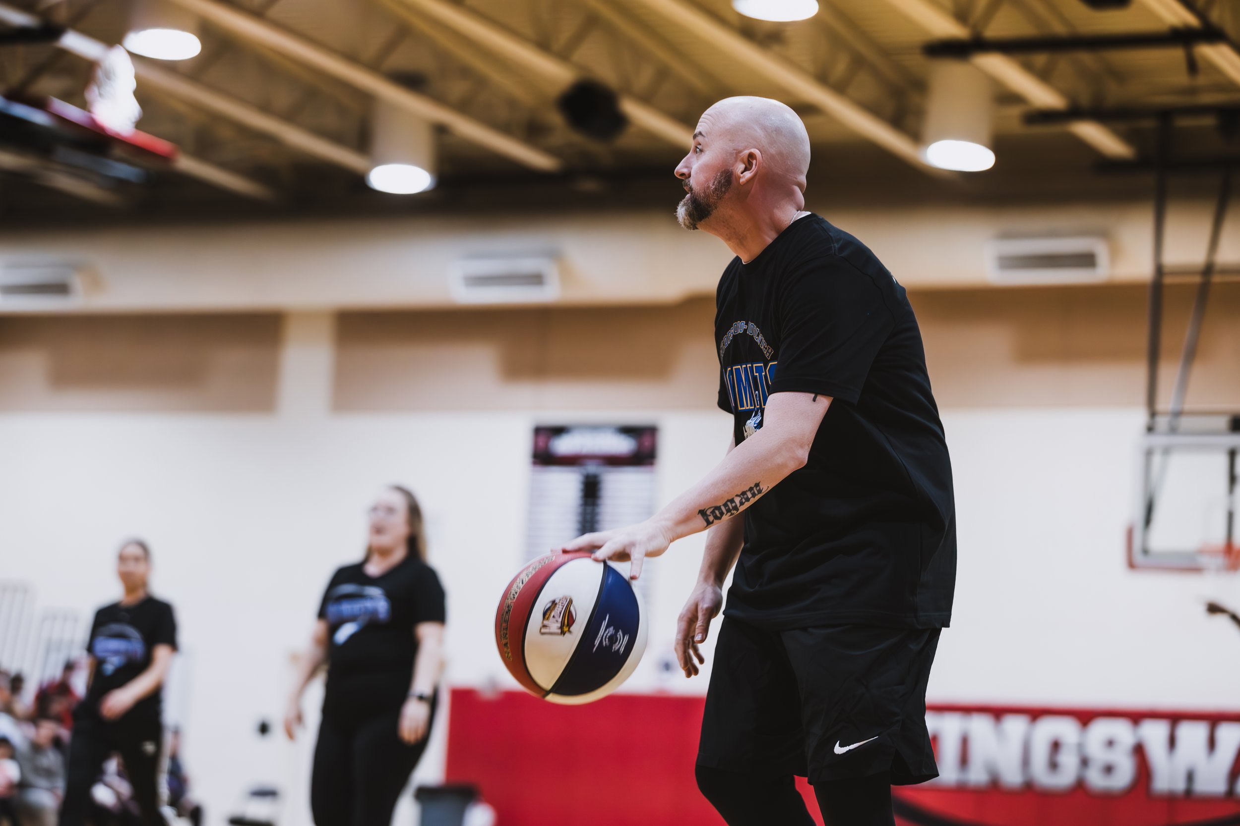 A man with a shaved head and beard playing basketball in a gym, holding a basketball, with two women blurred in the background.