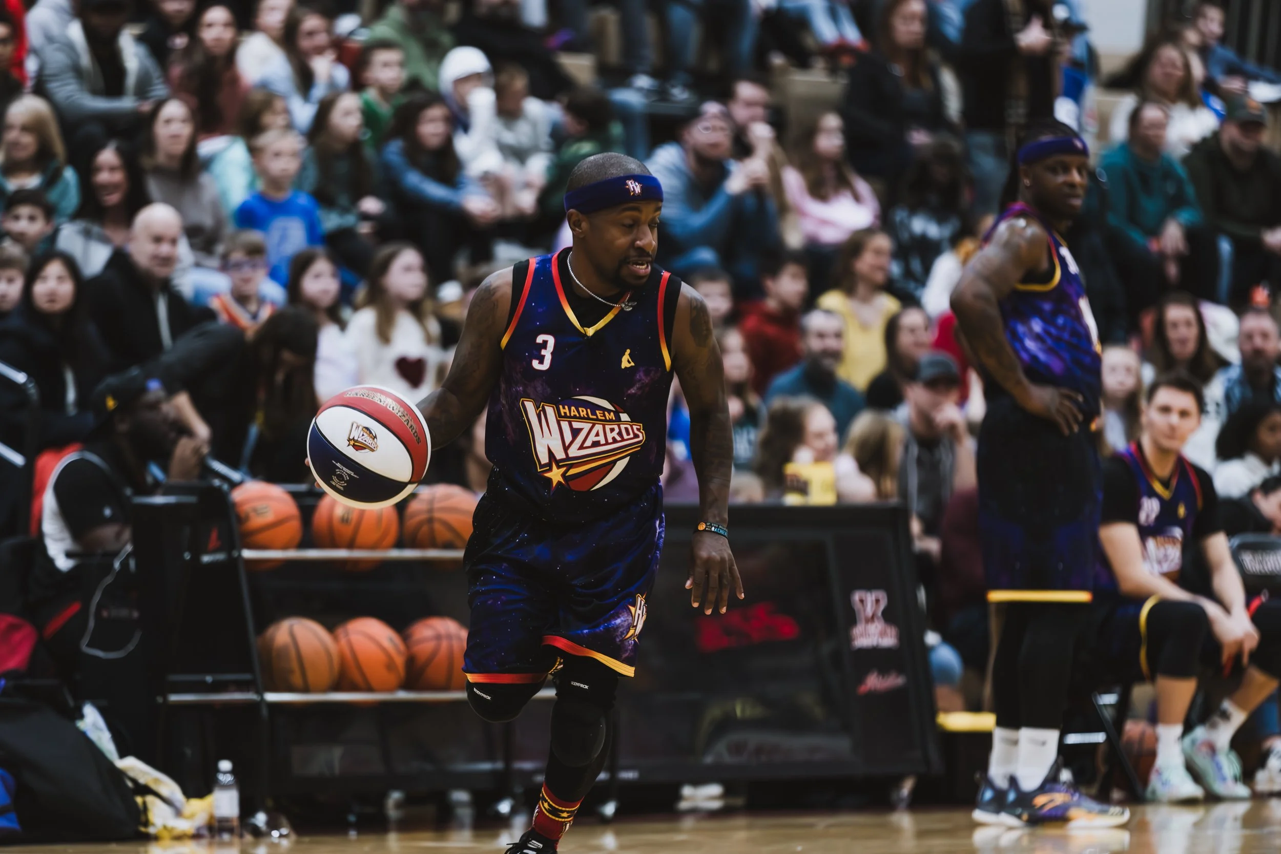 A basketball player from the Harlem Wizards, wearing a jersey with the number 3, dribbling a basketball on the court during a game, with spectators watching in the background.
