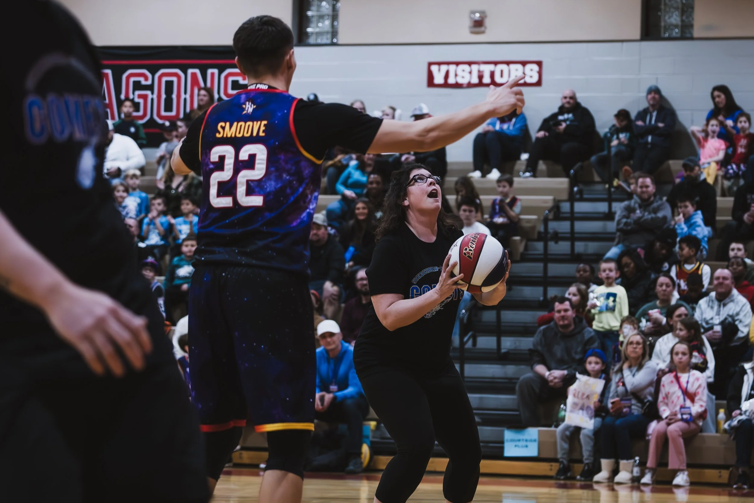 A woman holding a basketball preparing to make a shot, with a male teammate and spectators in the background at an indoor basketball game.