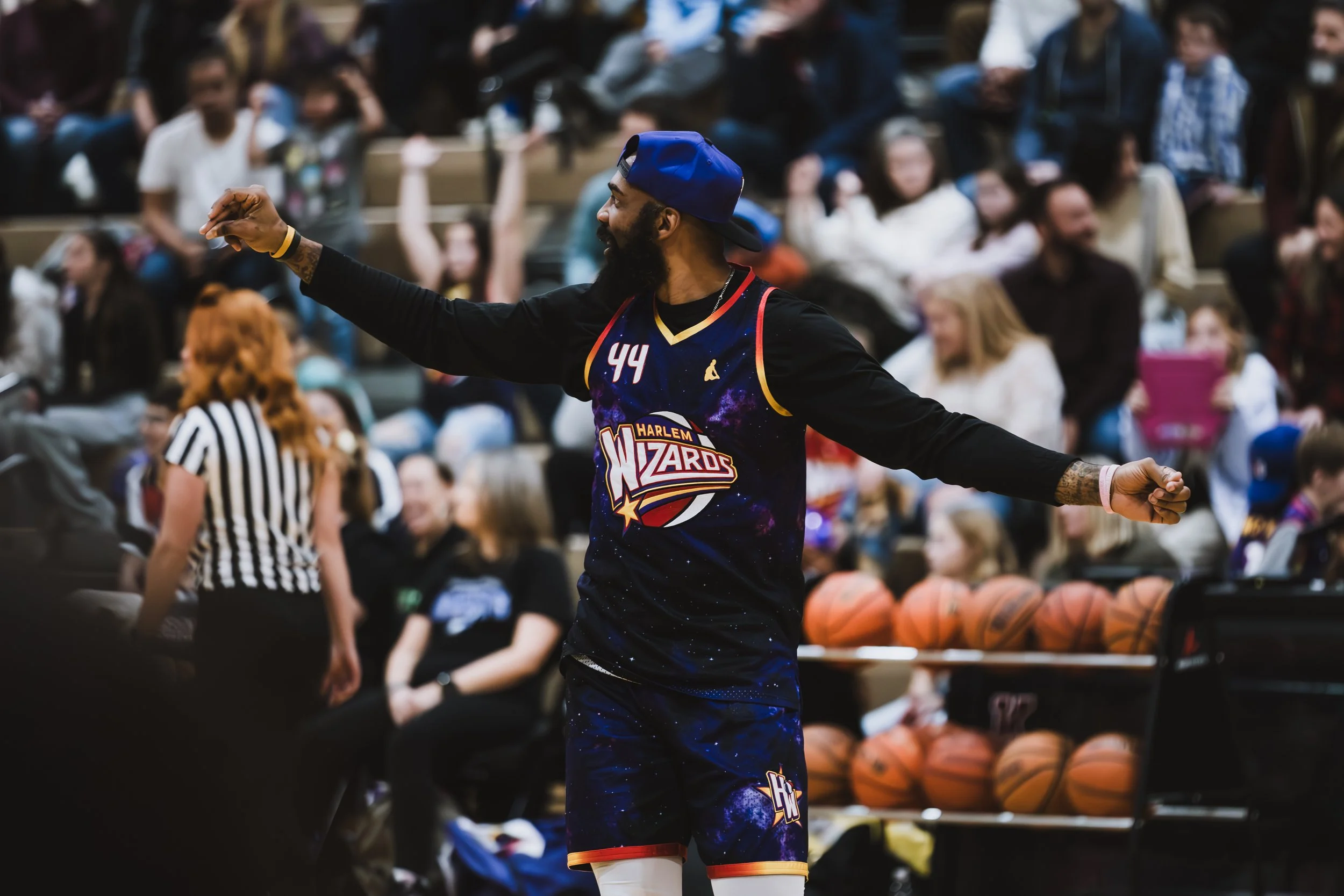 A basketball player in a Harlem Wizards uniform standing on the court with arms outstretched, surrounded by spectators and basketballs in the background.