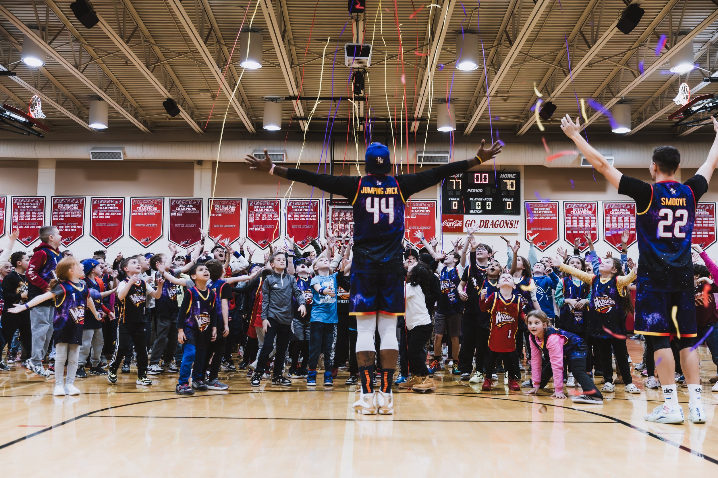 A basketball coach stands with arms outstretched in front of children and teenagers during a celebration event in an indoor gymnasium, with confetti and colorful streamers falling from the ceiling, and banners hanging on the wall.