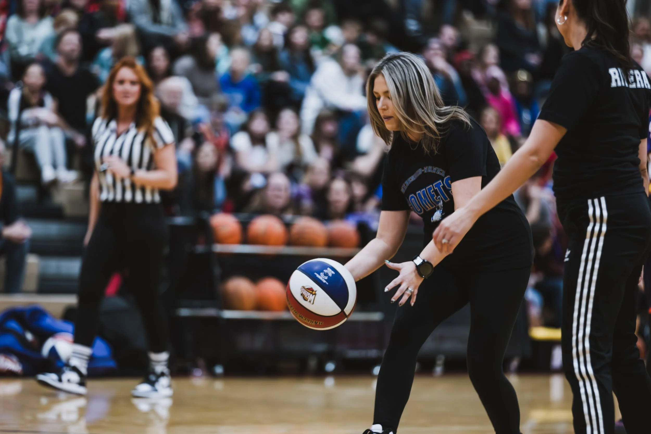 A female school staff member in a black event t-shirt smiling as she handles a red, white, and blue basketball during the PTO fundraiser game.