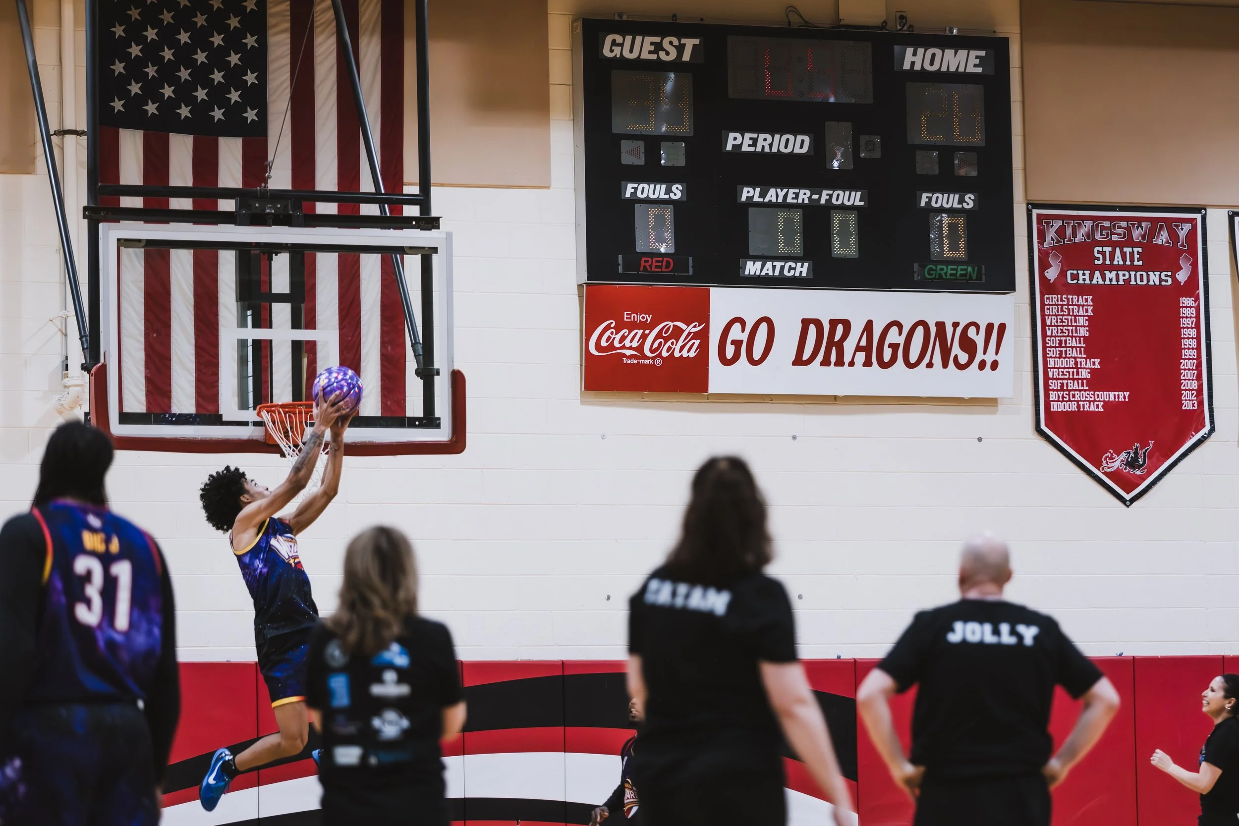 A wide shot of a player taking a shot toward the basket with the 'Go Dragons!!' school scoreboard and an American flag in the background.