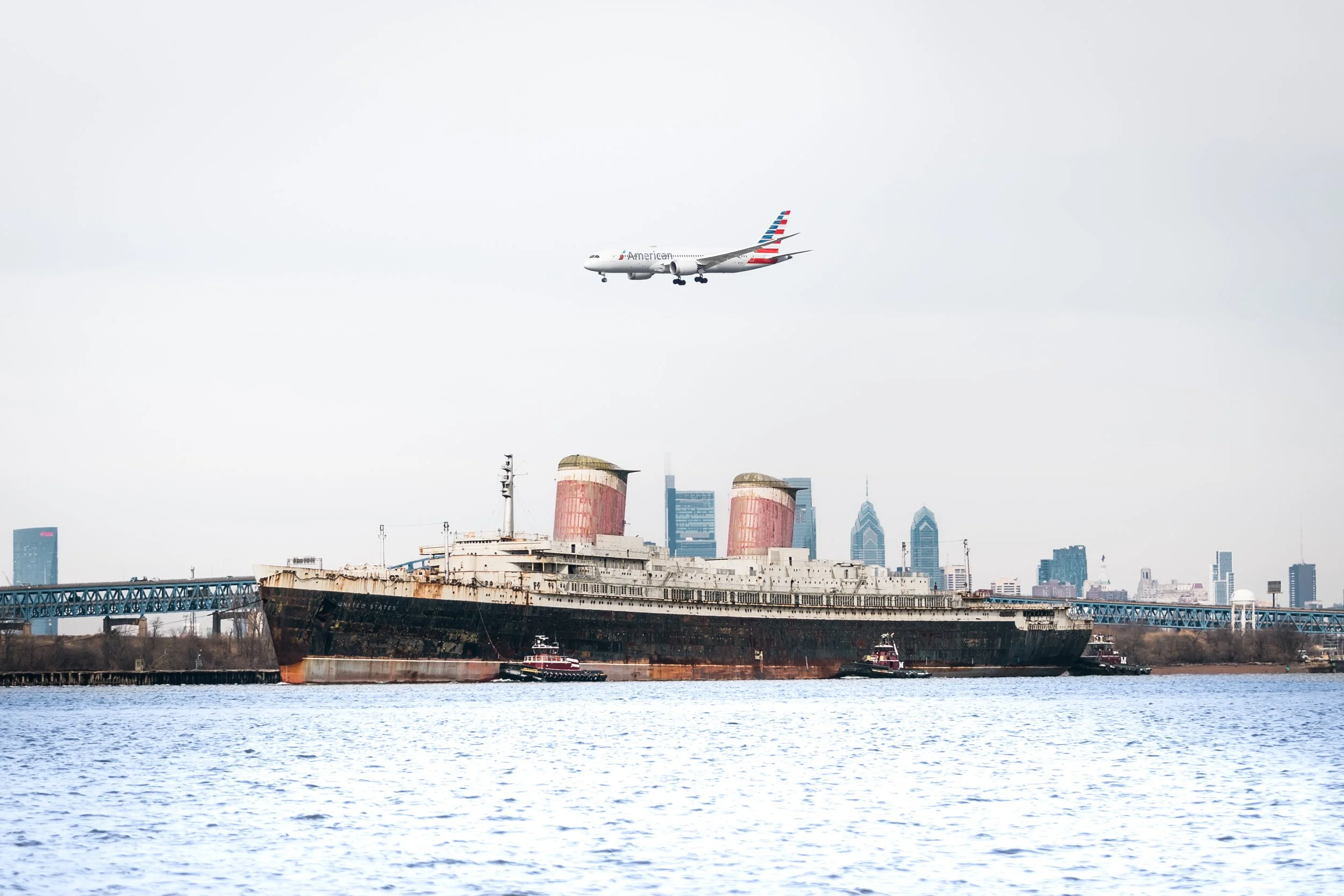 An American Airlines airplane flying over a large, abandoned shipwreck in the water with a city skyline in the background.