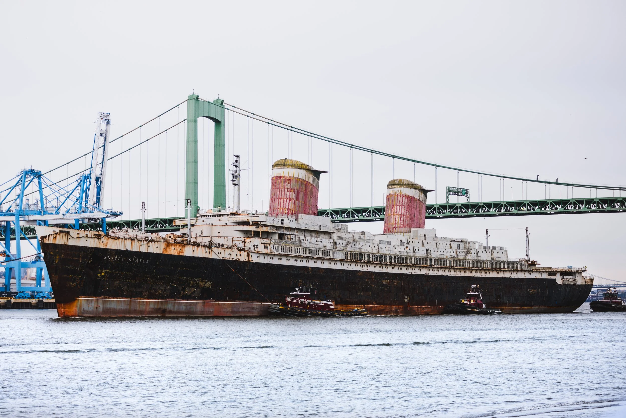 An old, rusty Titanic ship sculpture positioned in a harbor with a large suspension bridge in the background.