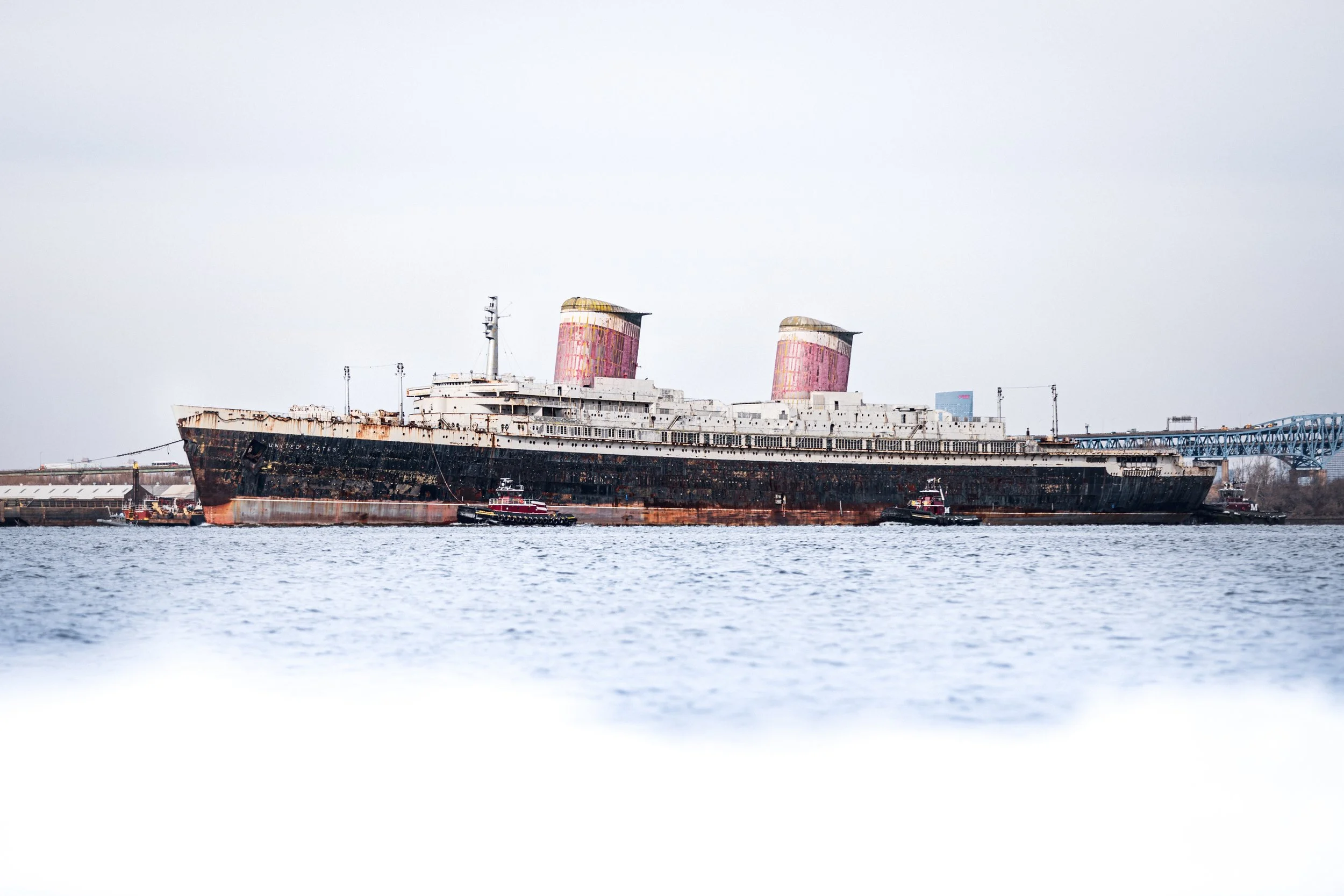 A large, rusted, abandoned cruise ship with two pink and yellow funnels docked at a port, with three smaller tugboats nearby and an overcast sky.