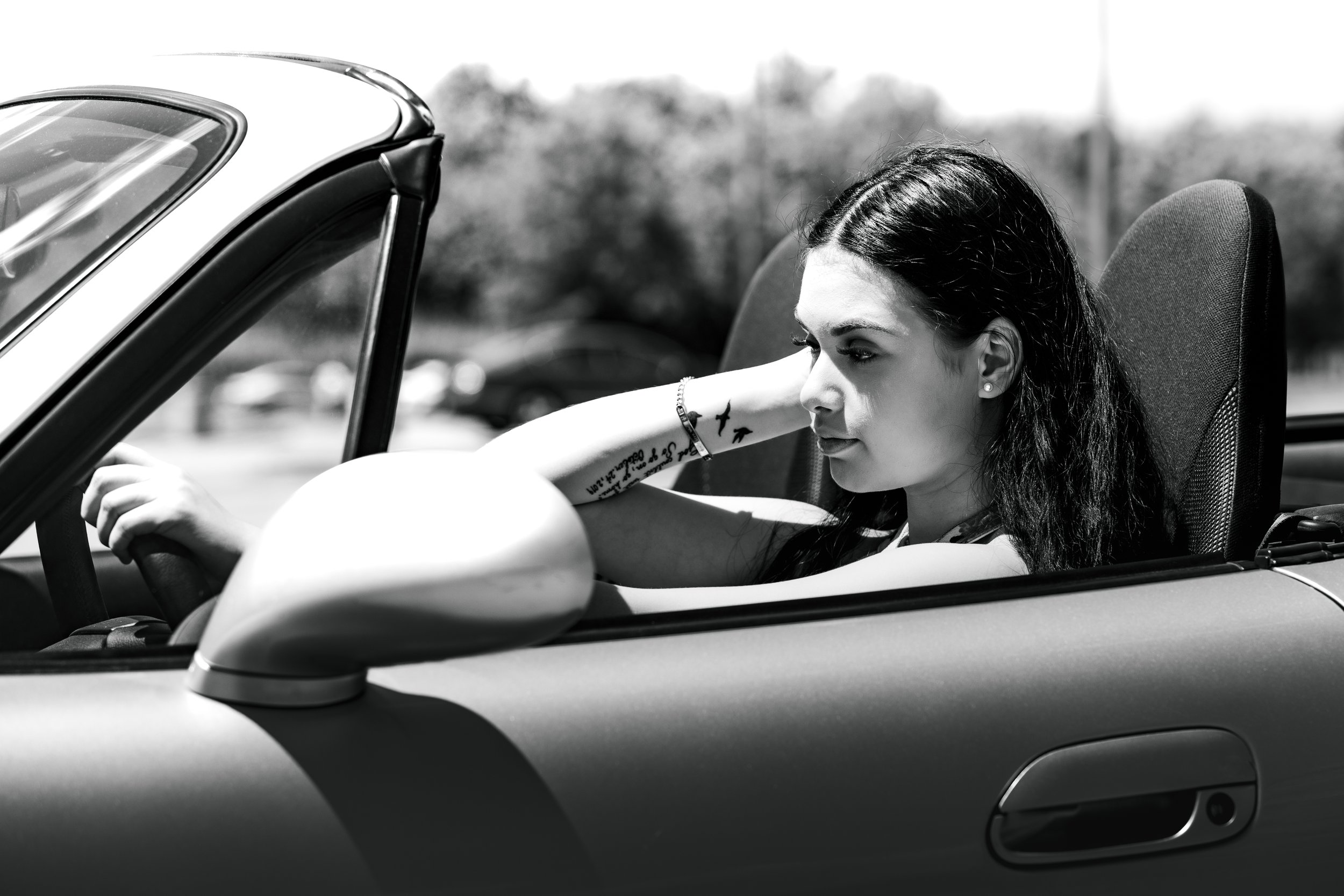 Black and white photo of a woman driving a convertible car, resting her arm on the door and looking to the side.