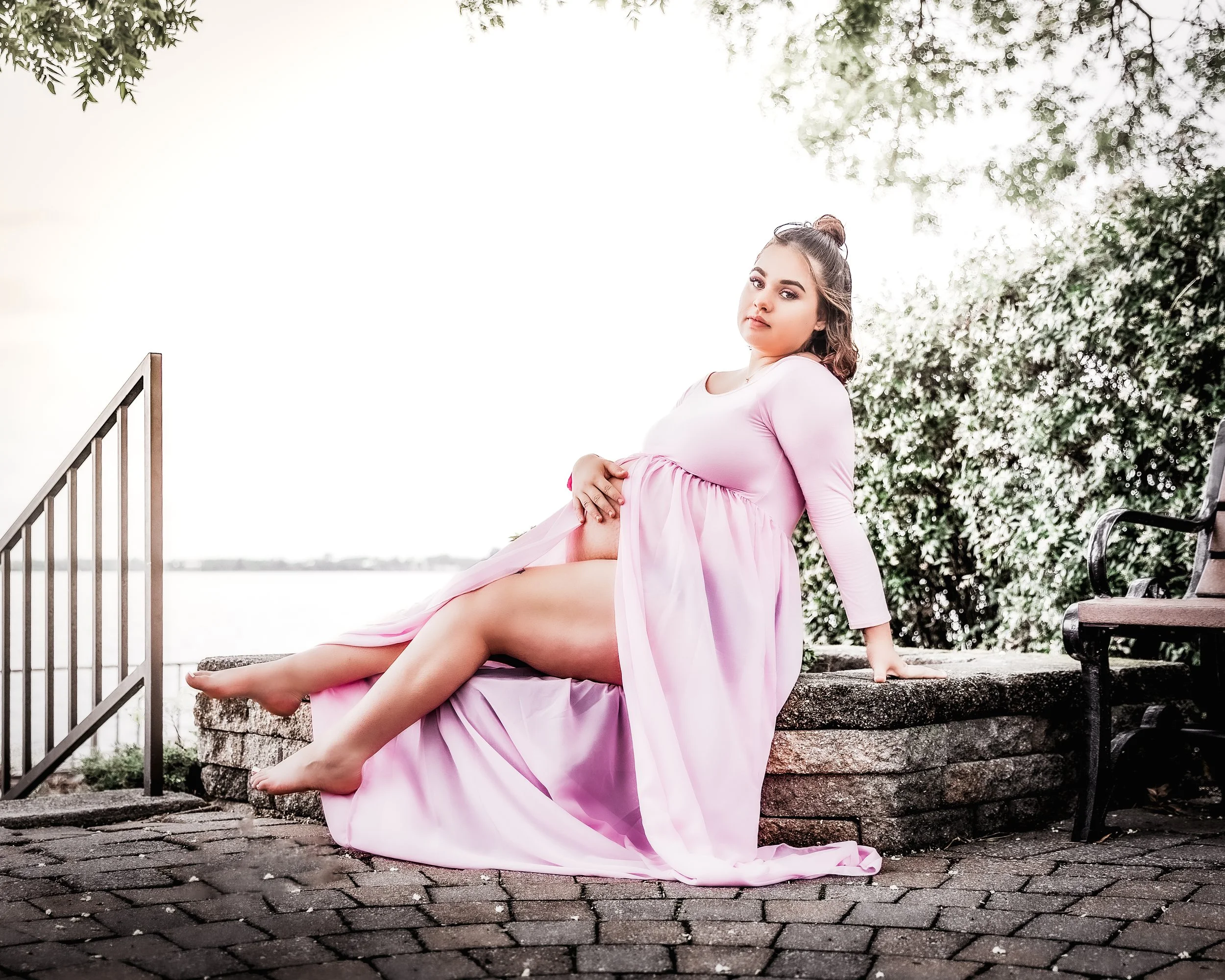 A pregnant woman in a pink dress sitting on a stone ledge outdoors near a body of water, with trees in the background. Maternity photography at Red Bank Battlefield in South Jersey. Photo by Digital Imageries Photography.