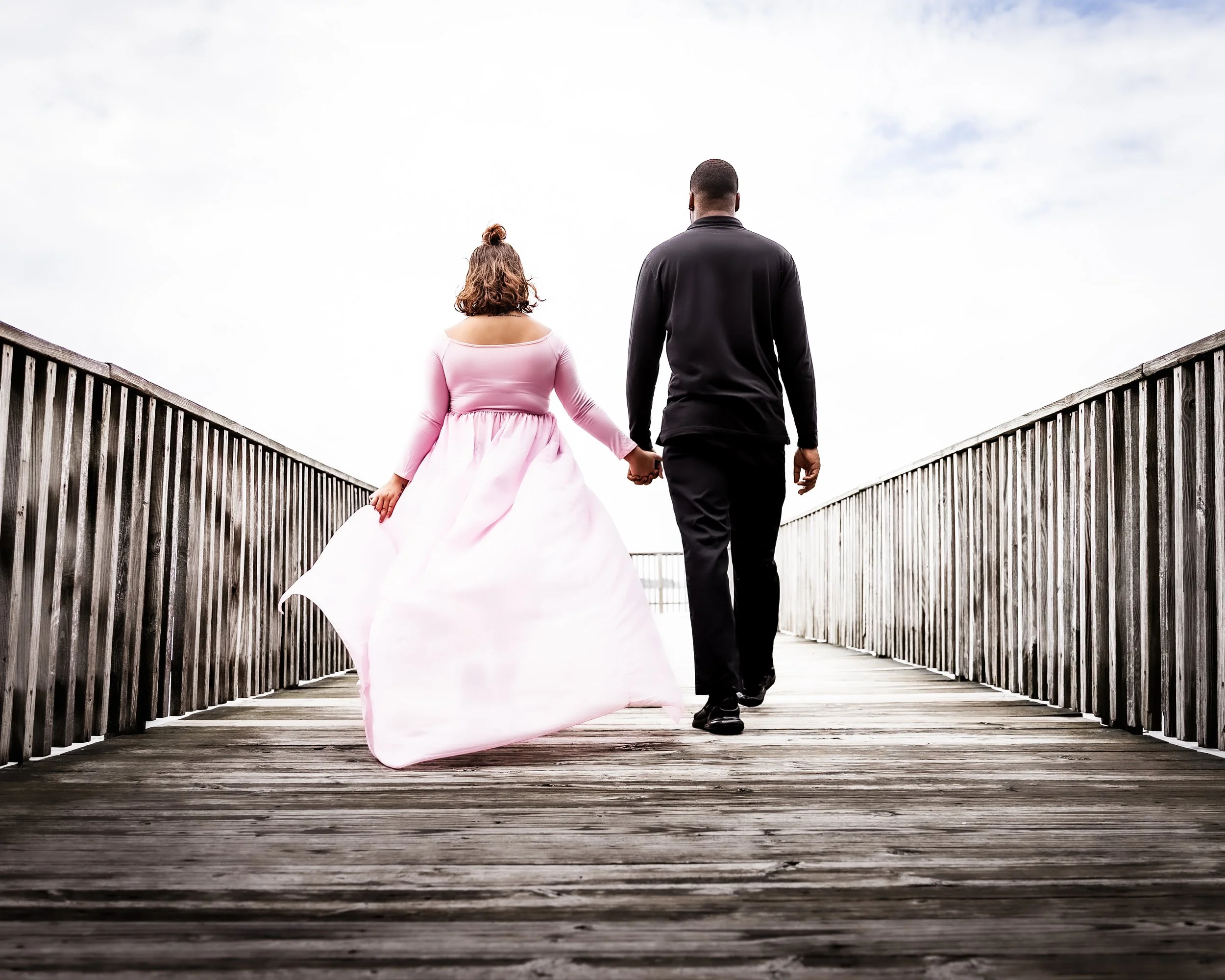 A couple walking hand in hand on a wooden bridge, the woman in a pink dress and the man in black.