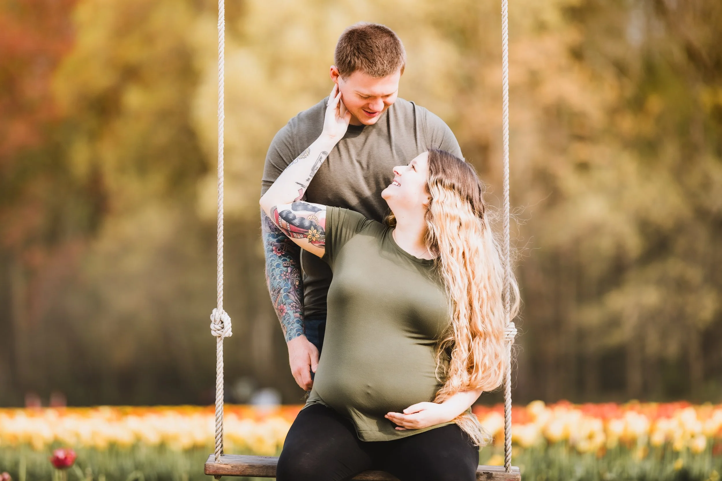 Pregnant woman with tattoos sitting on a swing, looking up and smiling at a man standing close behind her, who is touching her face gently, in a park with autumn trees and colorful flowers in the background.