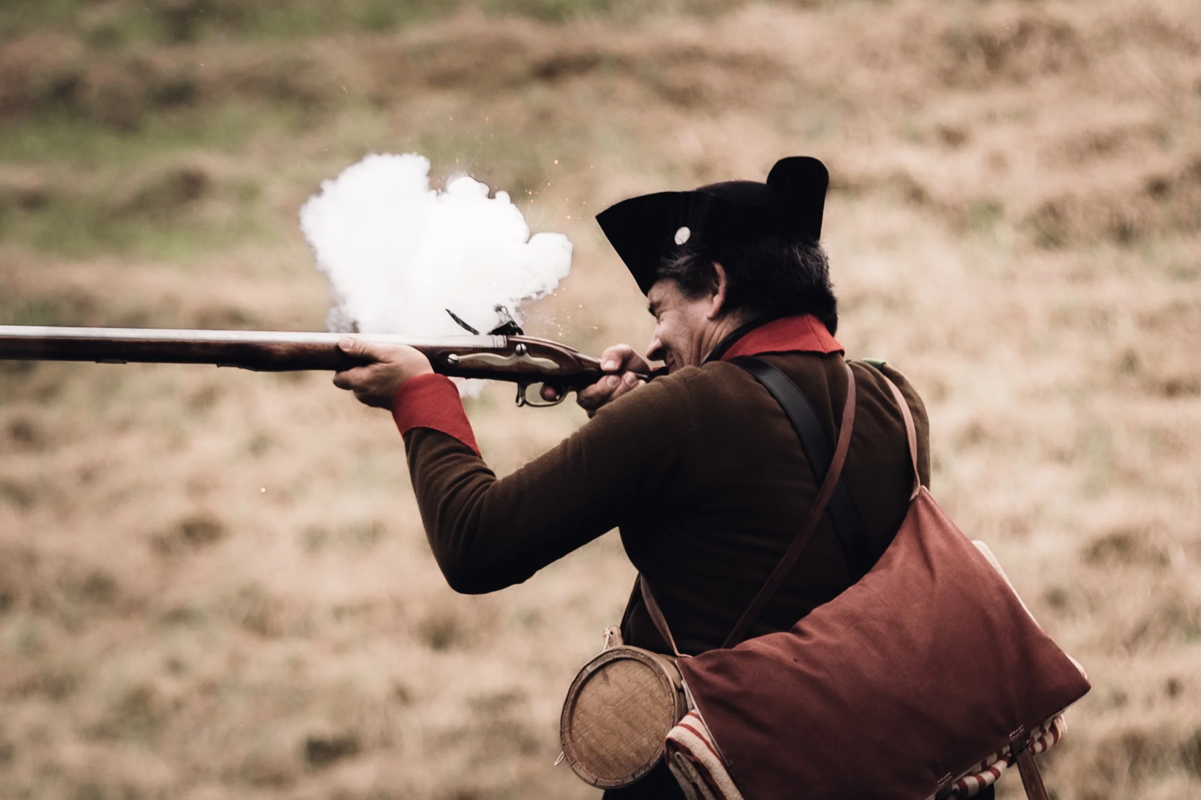 A reenactor dressed in 18th-century military uniform firing a musket, with smoke and spark coming from the barrel, located at Fort Mott in New Jersey.