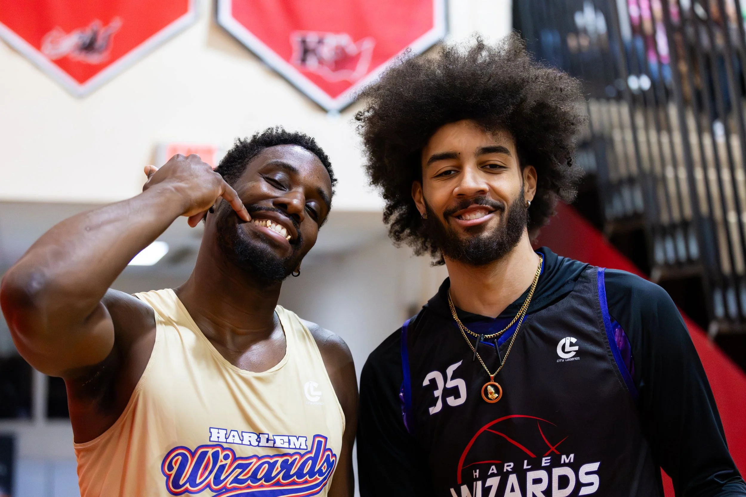 Two men smiling in a gym, one wearing a Harlem Wizards basketball jersey and the other in a black Lakers basketball jersey. They are standing close together with a background of sports banners and gym equipment.