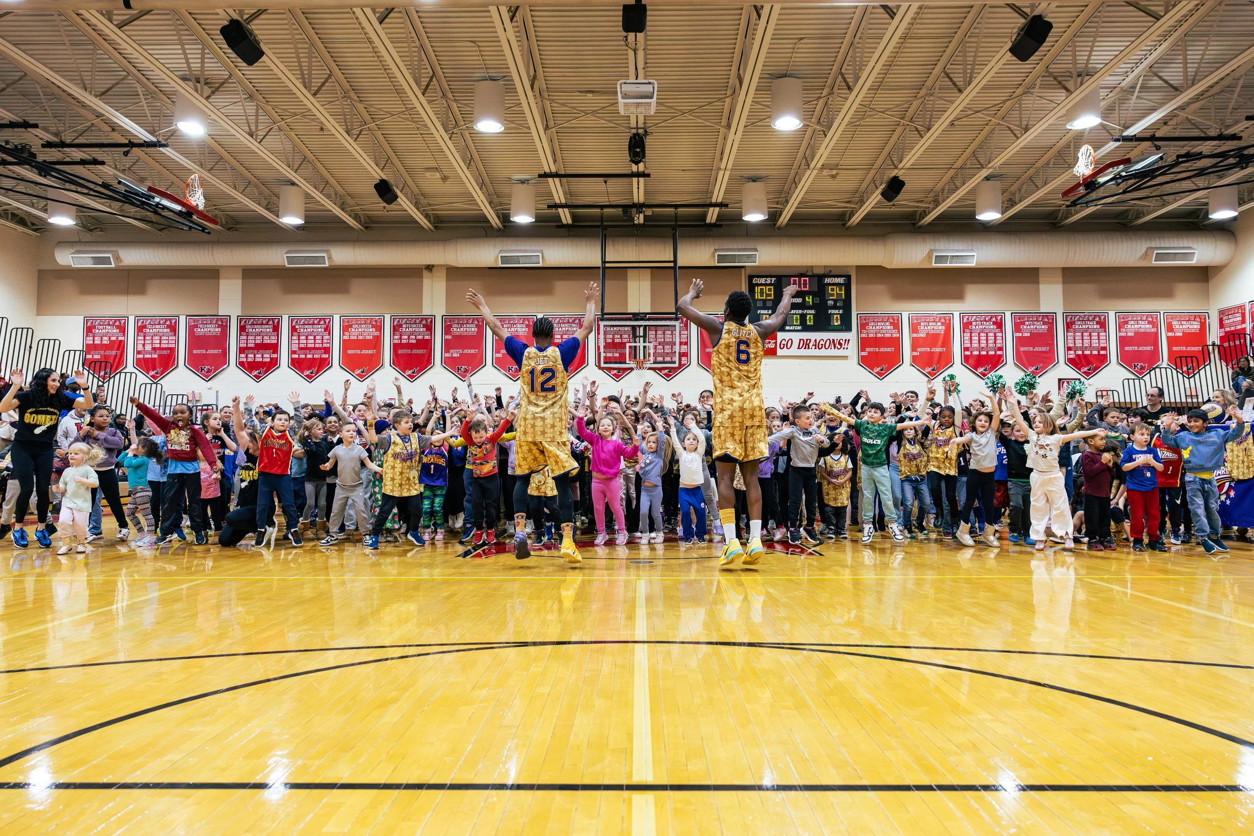 Basketball players in yellow uniforms leading a dance or exercise with a large group of children and adults in a gymnasium with banners on the wall.