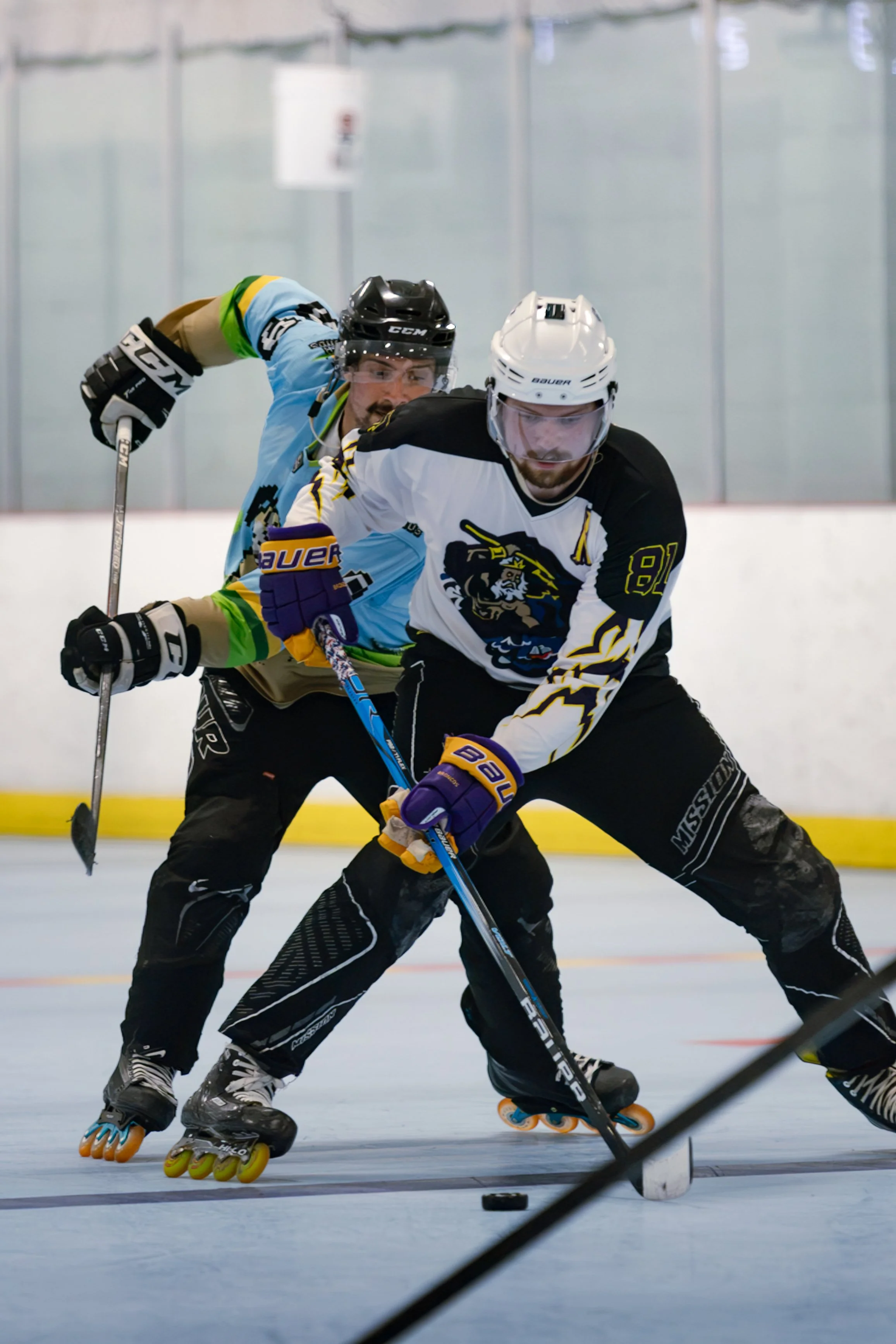 Two roller hockey players in motion on the rink, one in black and white with a blonde beard and the other in a blue and green jersey, both wearing helmets and gloves, competing for the puck.