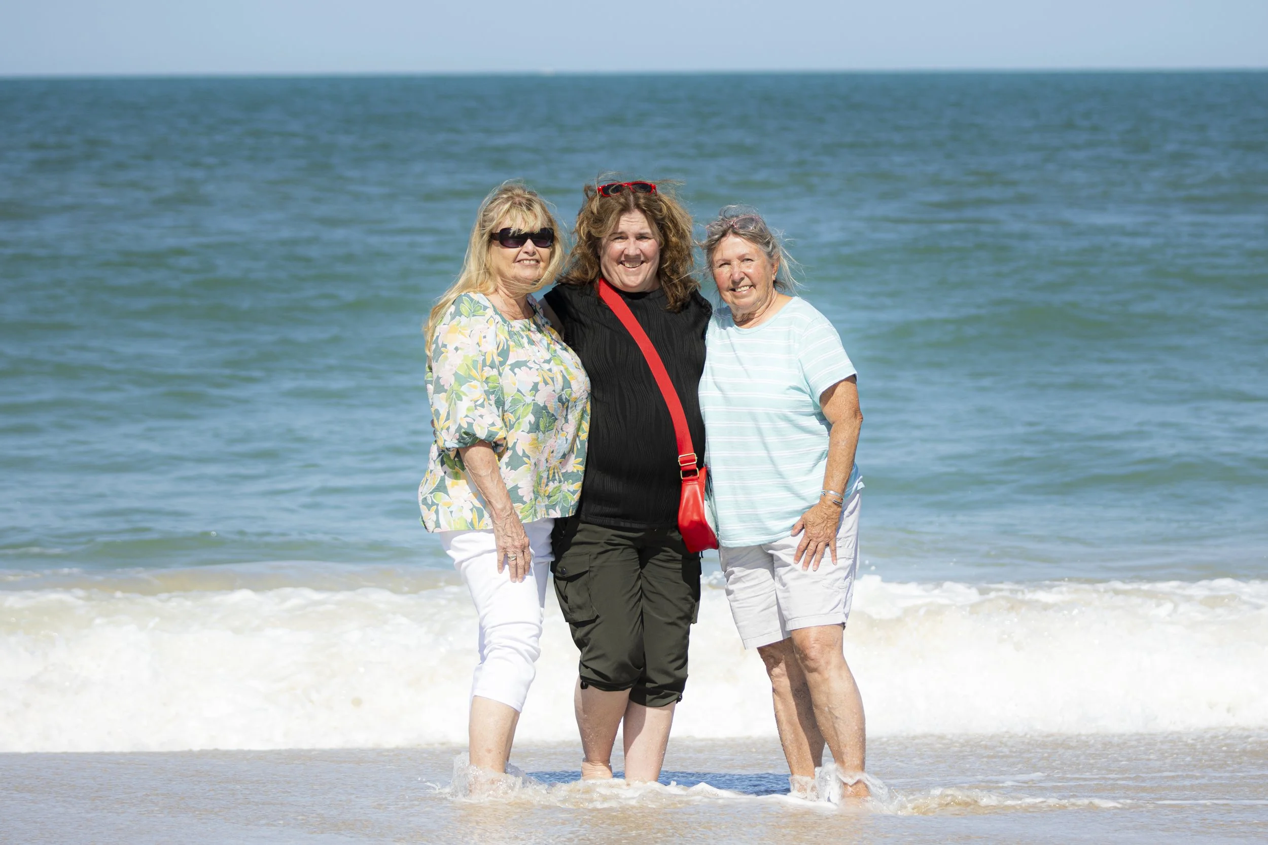 Three women standing close together at the beach with ocean waves in the background, smiling for the camera.