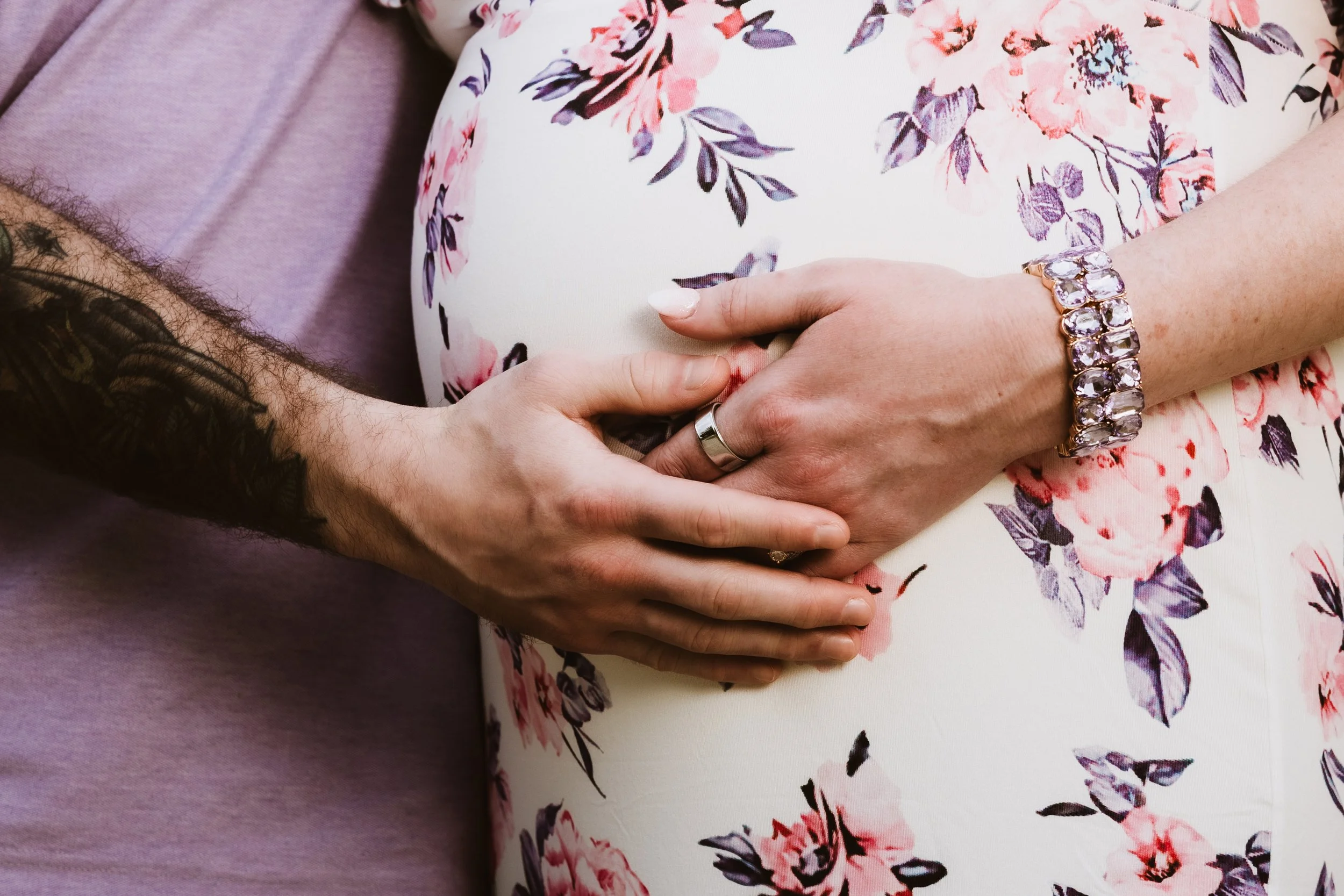 A couple's hands are intertwined, with the woman wearing a large ring and a bracelet with stones and the man wearing a plain silver wedding band. The woman's floral dress is visible, and the man's sleeve is purple, showcasing a tattoo on his arm.