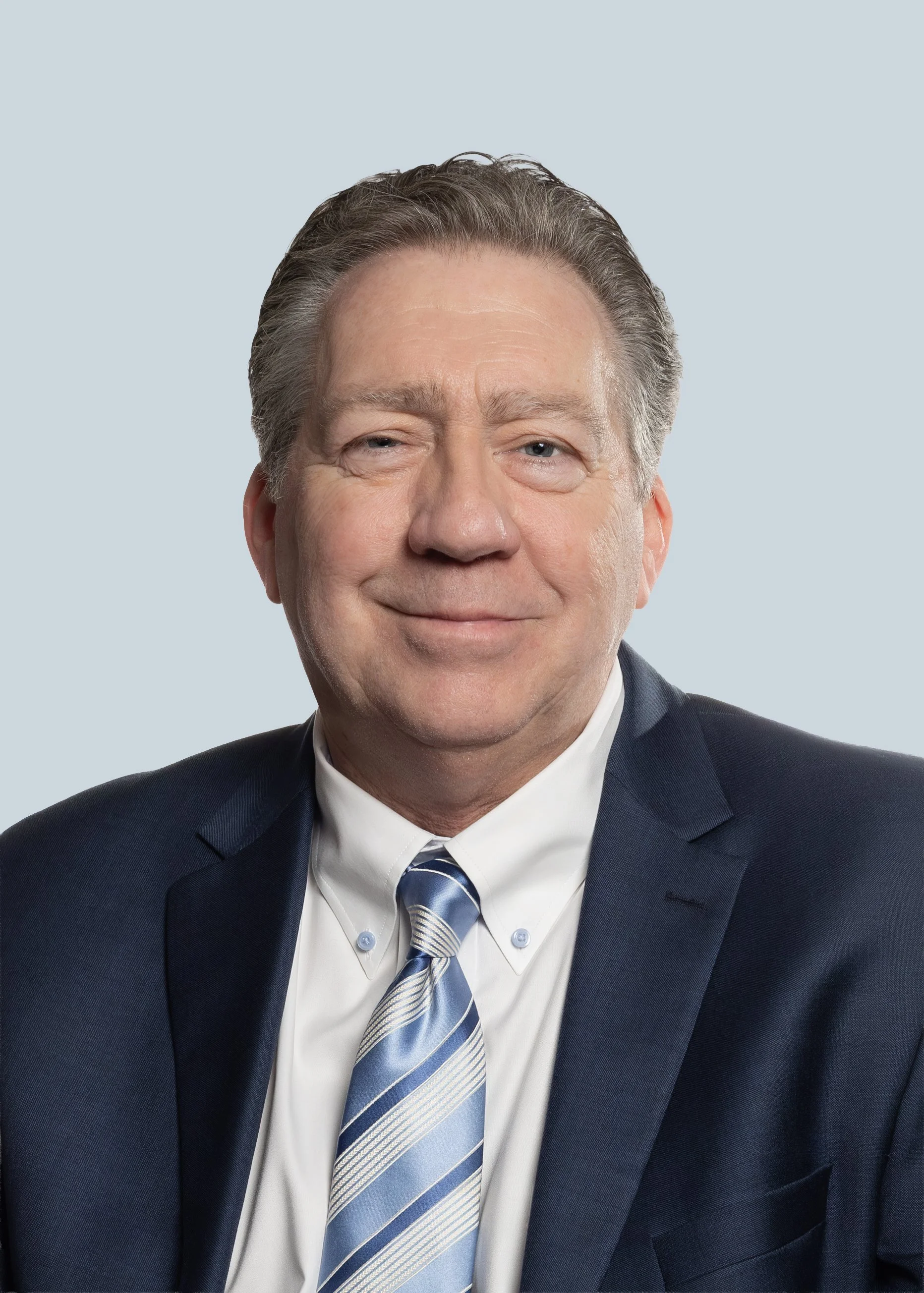 A middle-aged man smiling wearing a dark suit, white shirt, and a blue striped tie, against a light gray background. Ready for his professional headshot photos to be taken.