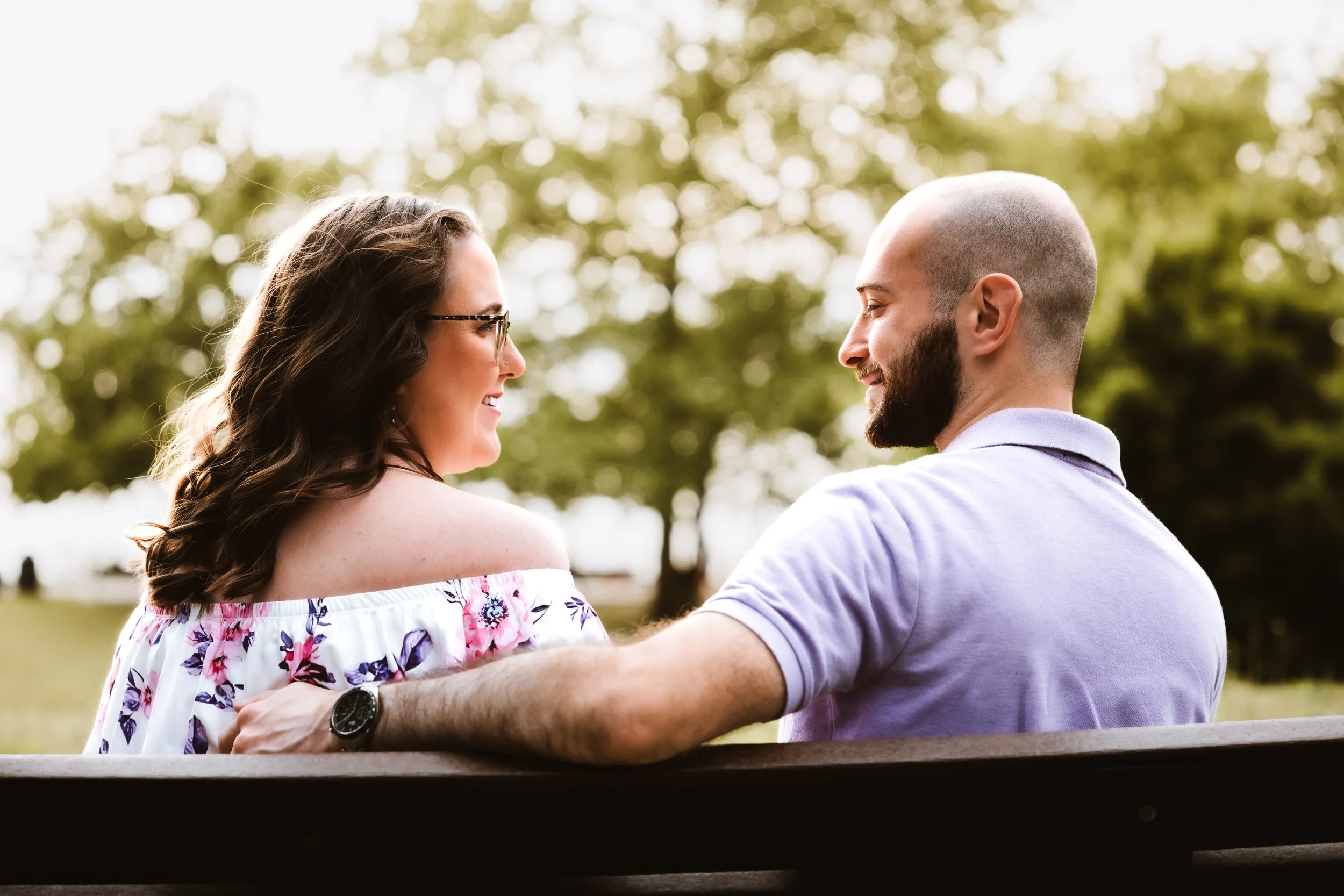 A man and woman face each other smiling, holding each other's arms, outdoors with trees in the background.