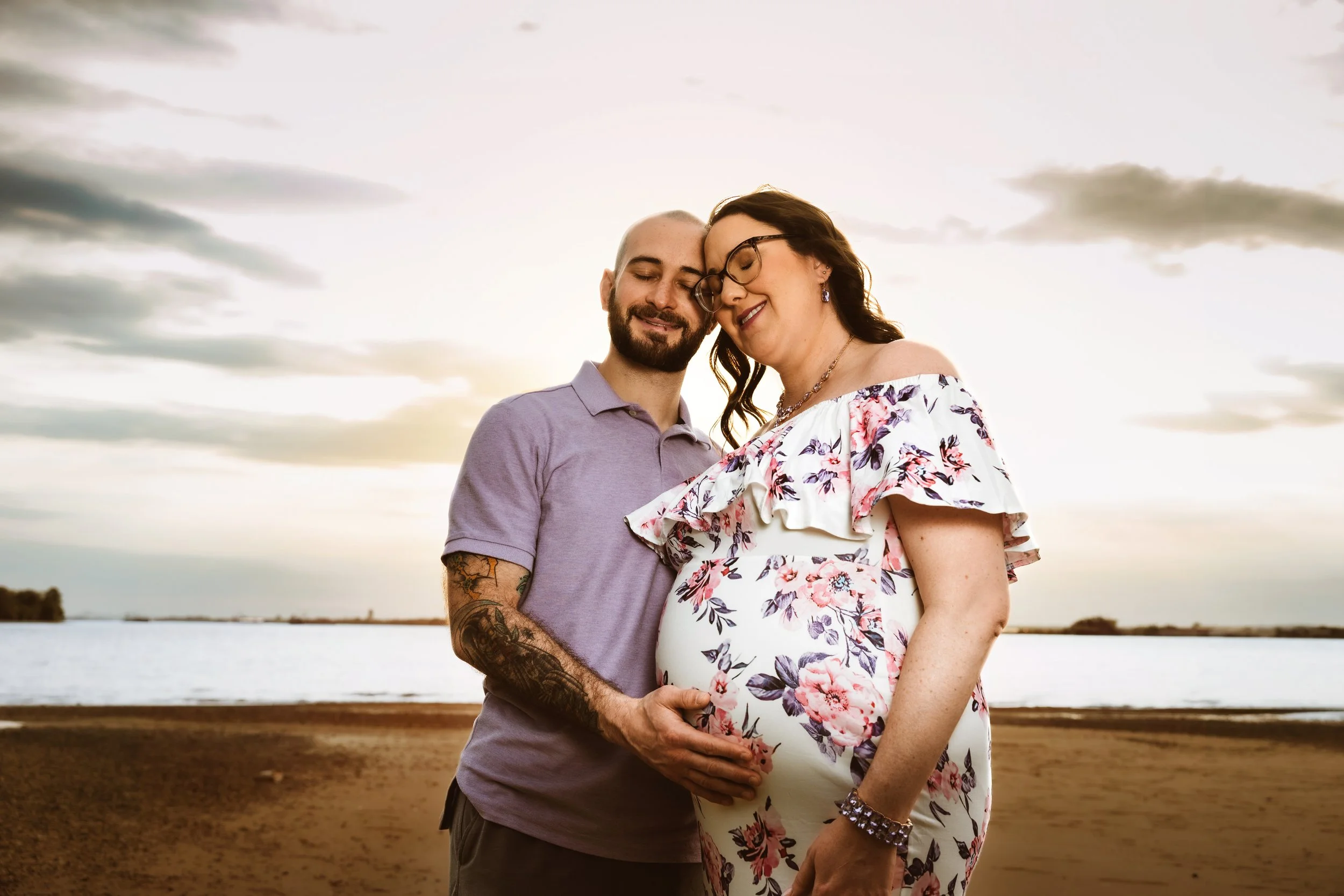 A pregnant woman wearing a floral dress and glasses and a man with a beard, a shaved head, and tattoos, wearing a purple shirt, are standing close together with their eyes closed, smiling, and touching their heads and belly on a beach during sunset.