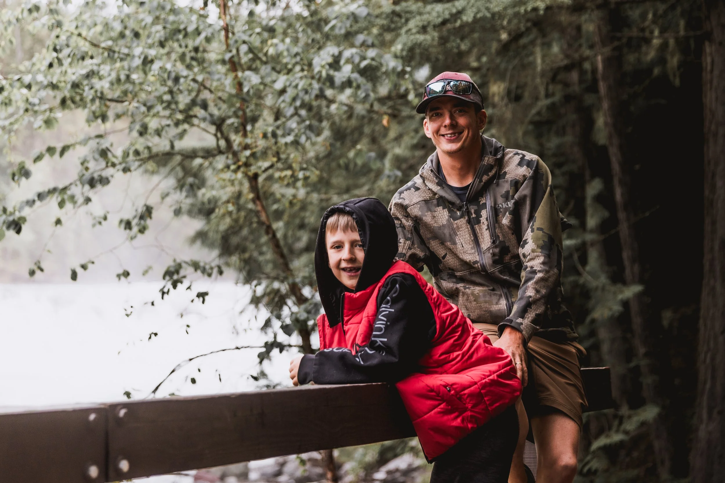 A man and boy smiling outdoors near a lake, with the man sitting on a wooden railing and the boy leaning on it, surrounded by trees.