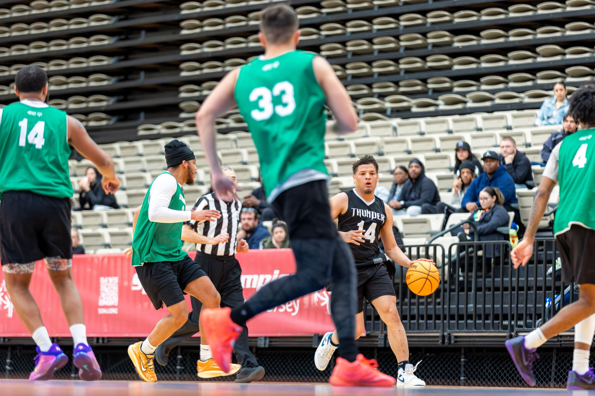 Basketball game with players on the court, one player in black dribbling, others in green jerseys, referees, and spectators in the background.