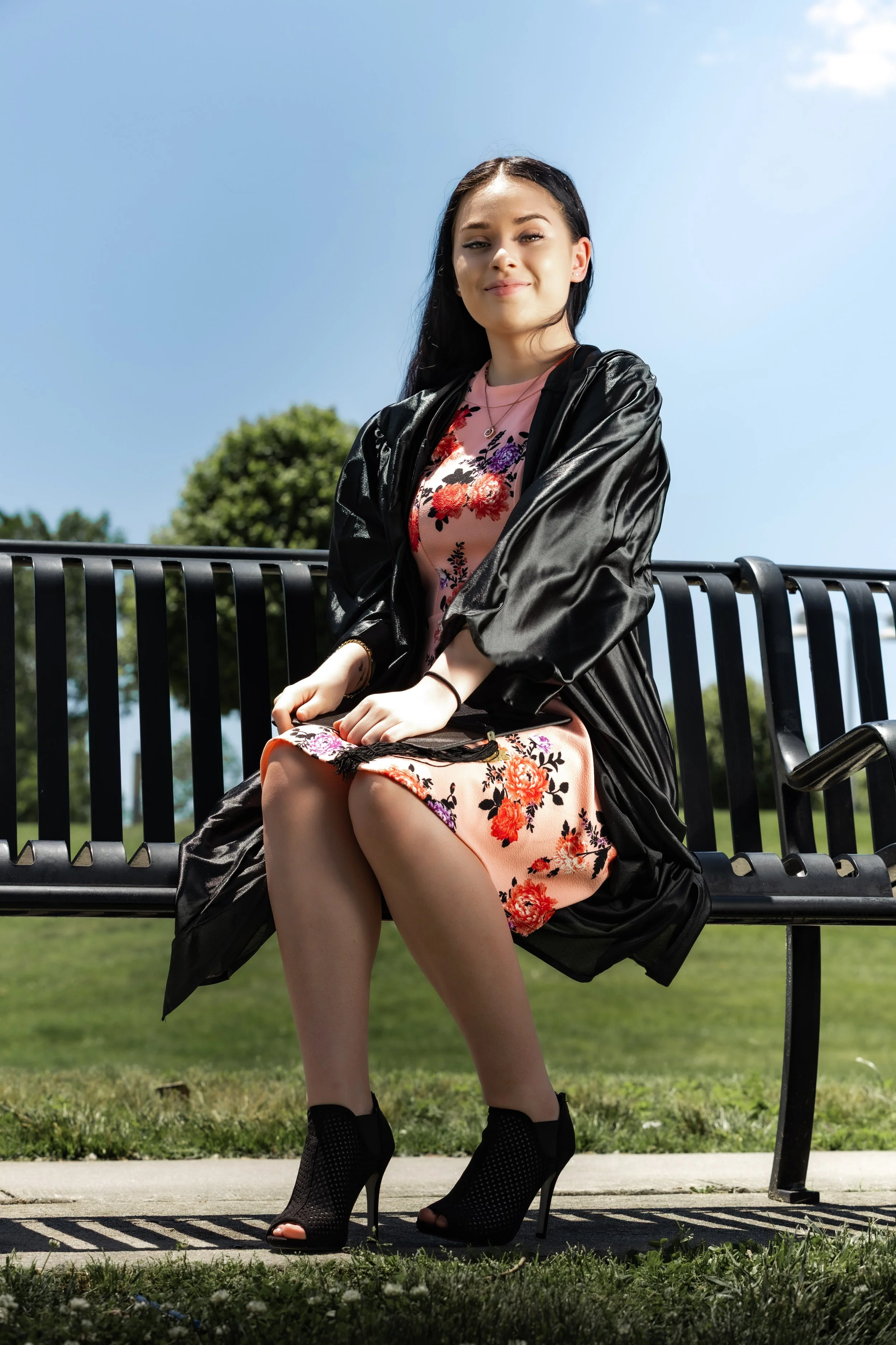A young woman with long dark hair sitting on a park bench, wearing a floral dress, black high heels, and a black leather jacket, smiling at the camera with a green park background and a clear blue sky.