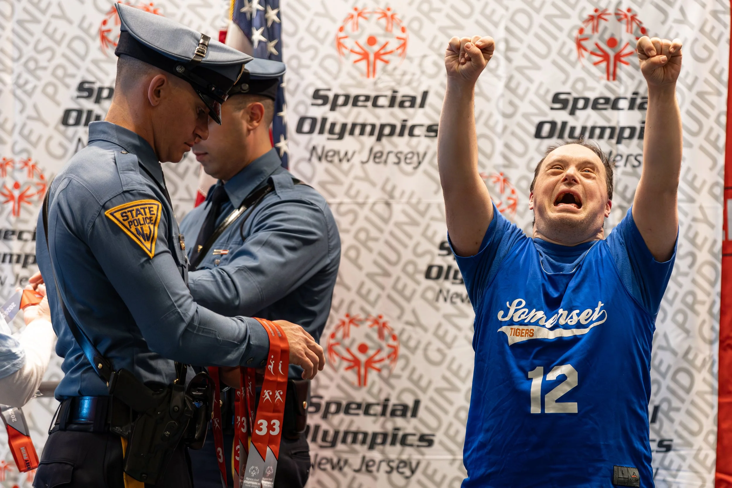 A man in a blue Homestest Tigers sports shirt with the number 12 raises both fists in celebration, with tears and an emotional expression on his face. Two uniformed police officers, one with a State Police patch, are nearby, holding medals. A backdro