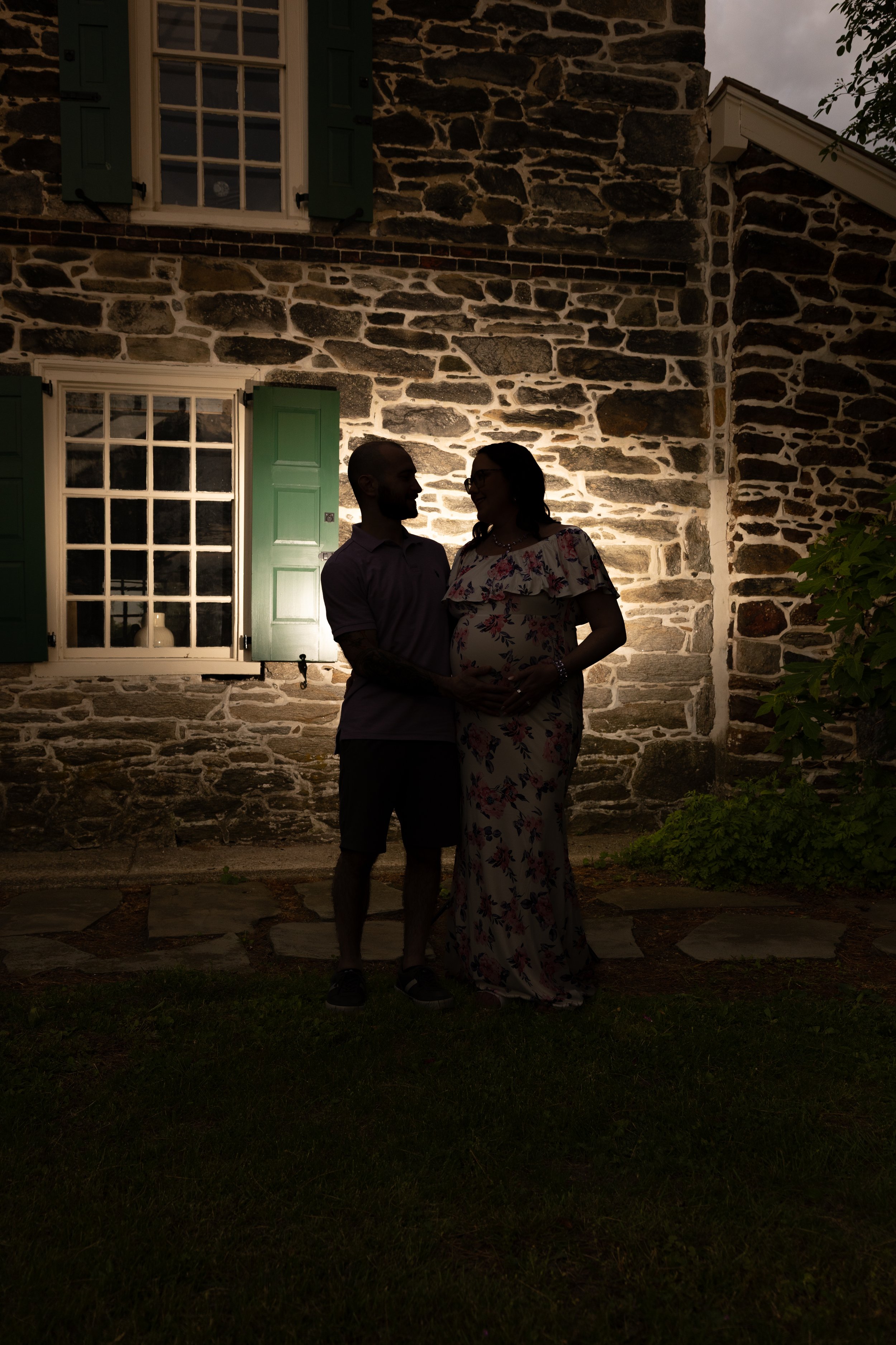 A couple stands close together outside at night, silhouetted against a stone house with lit windows and open green shutters.