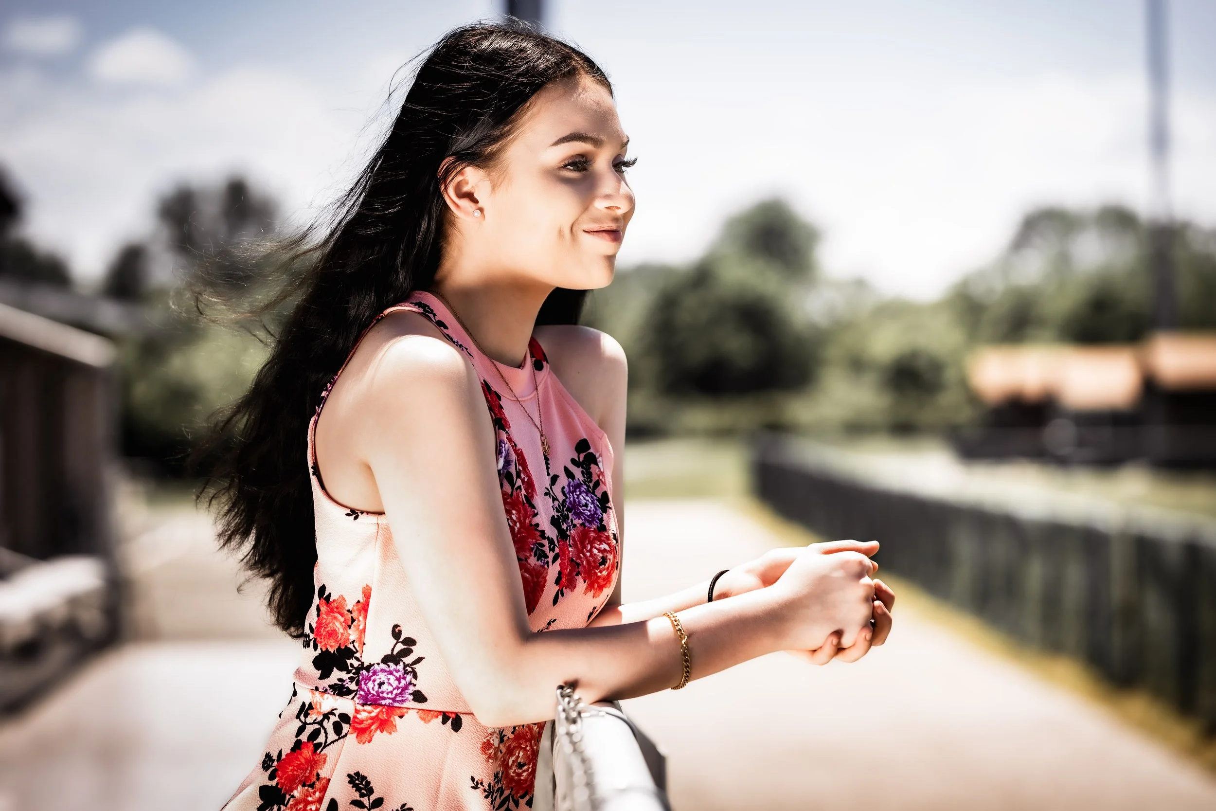 Young woman with dark hair wearing a pink floral dress smiling outdoors on a sunny day.