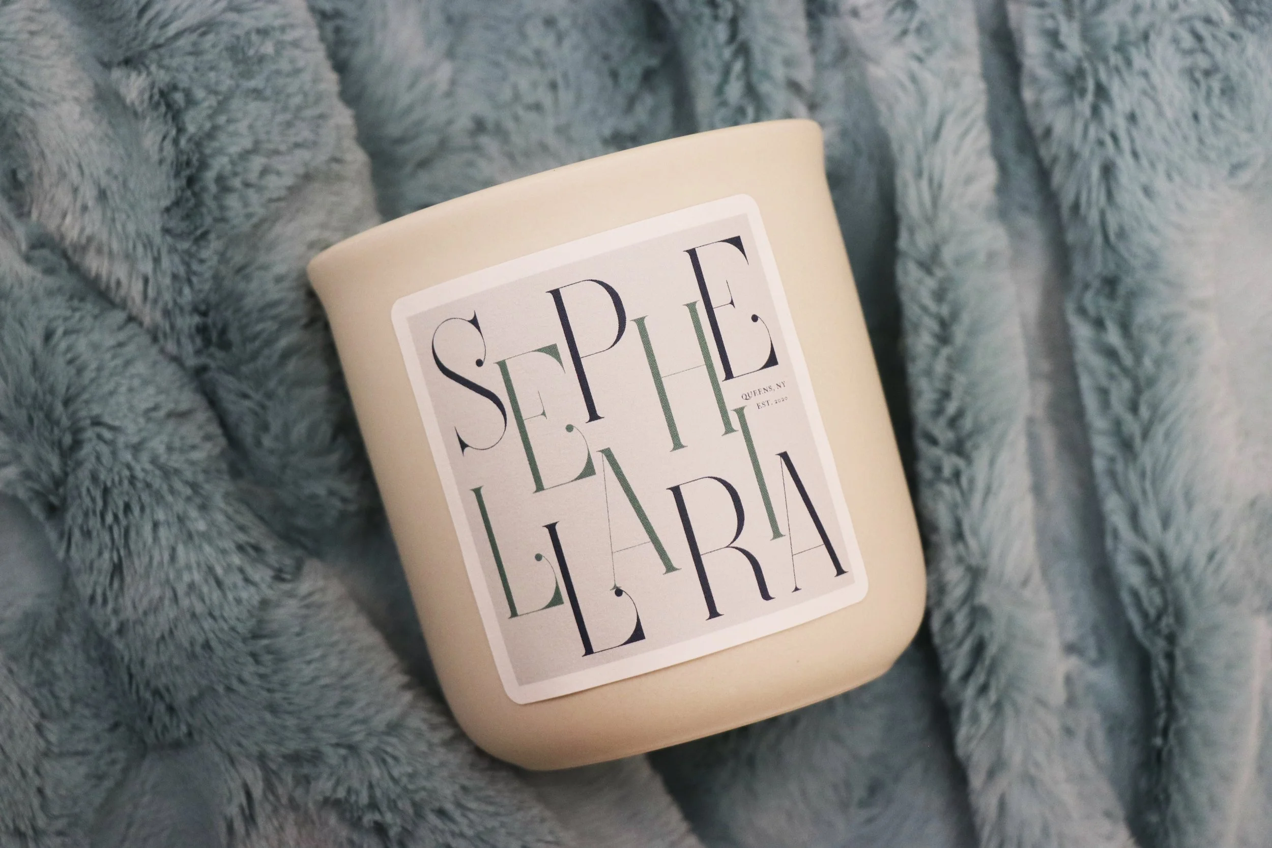 A beige scented candle with a label that reads 'SEPTEMBER' placed on a soft, gray faux fur blanket.