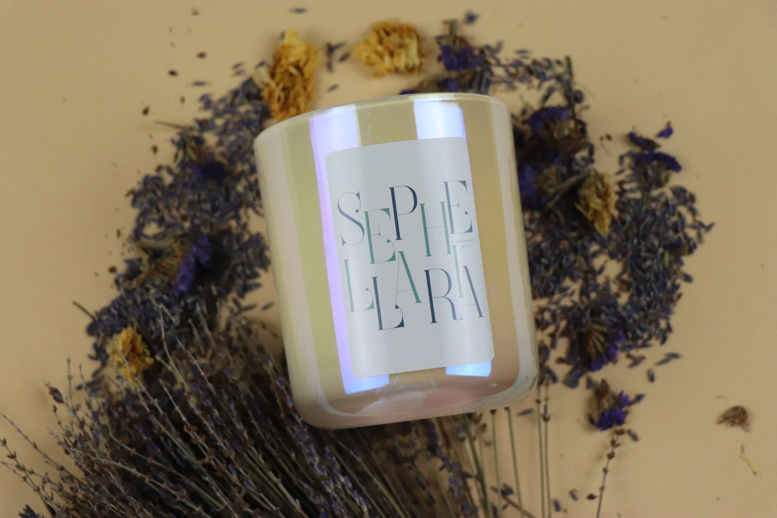 A white candle with a small piece of wood on top, surrounded by green pine branches, placed near a beige decorative object and a green container, against a light-colored wall.