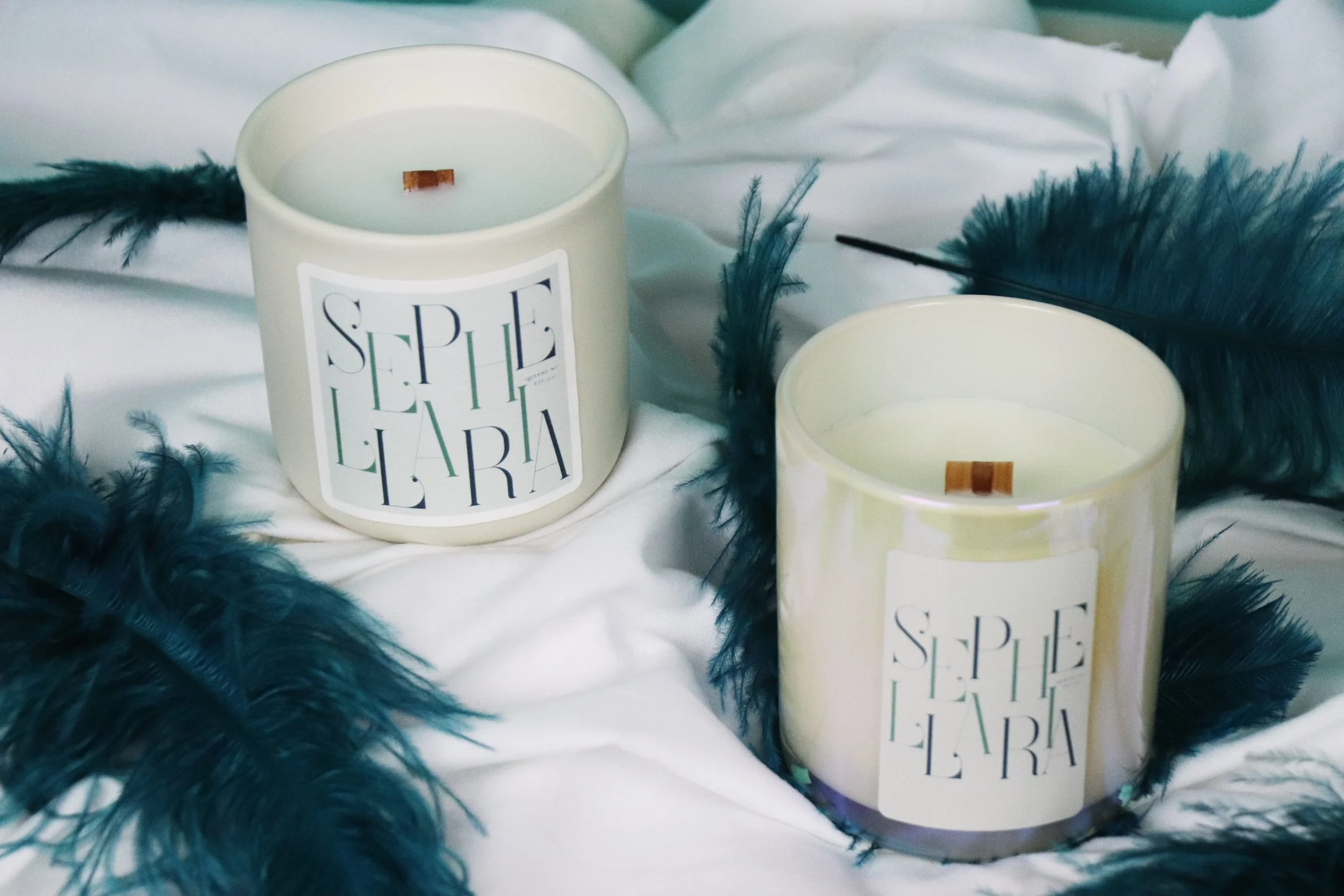 Two white scented candles with gold accents and labels that read 'SPELLE LAURA', surrounded by dark teal feathers on white fabric.