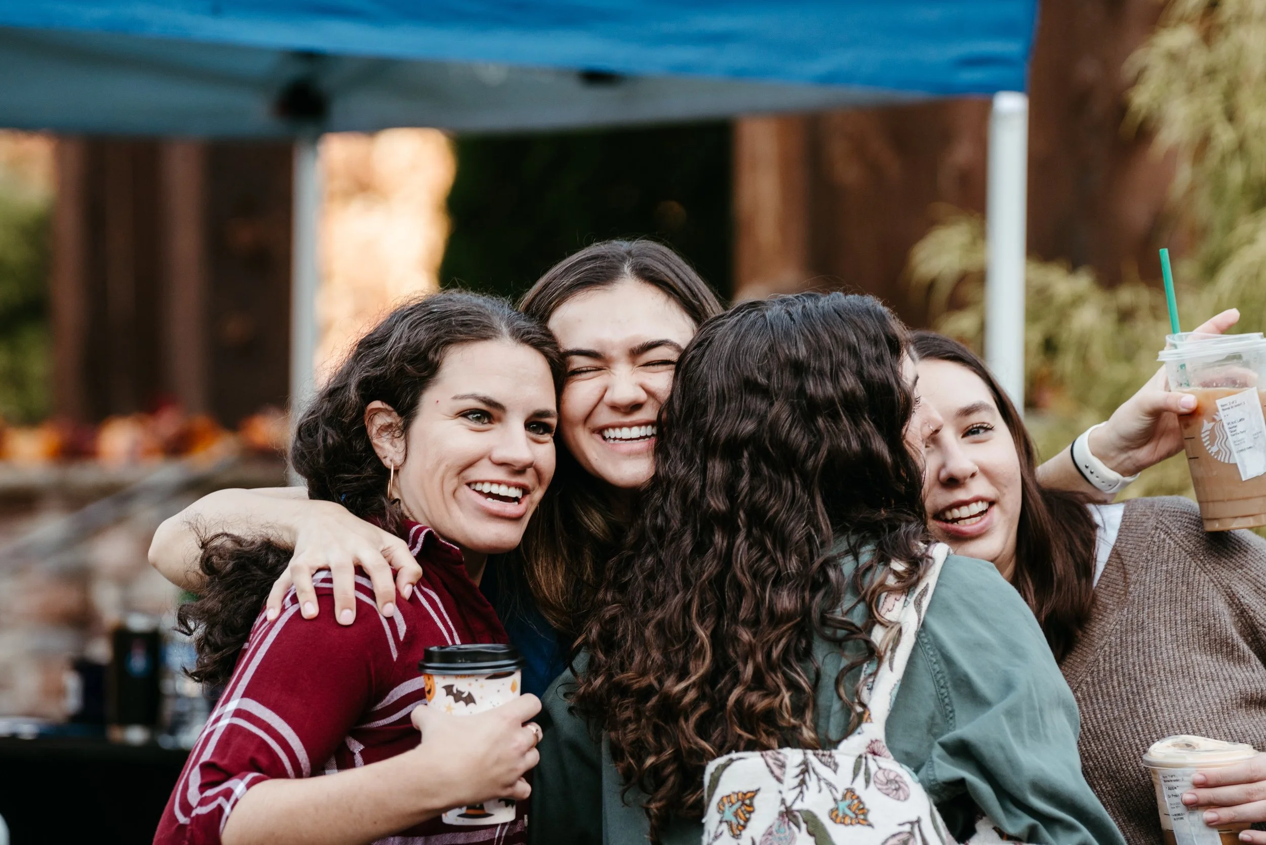 A group of diverse young women smiling and hugging outdoors during autumn, holding coffee cups and wearing casual clothes.
