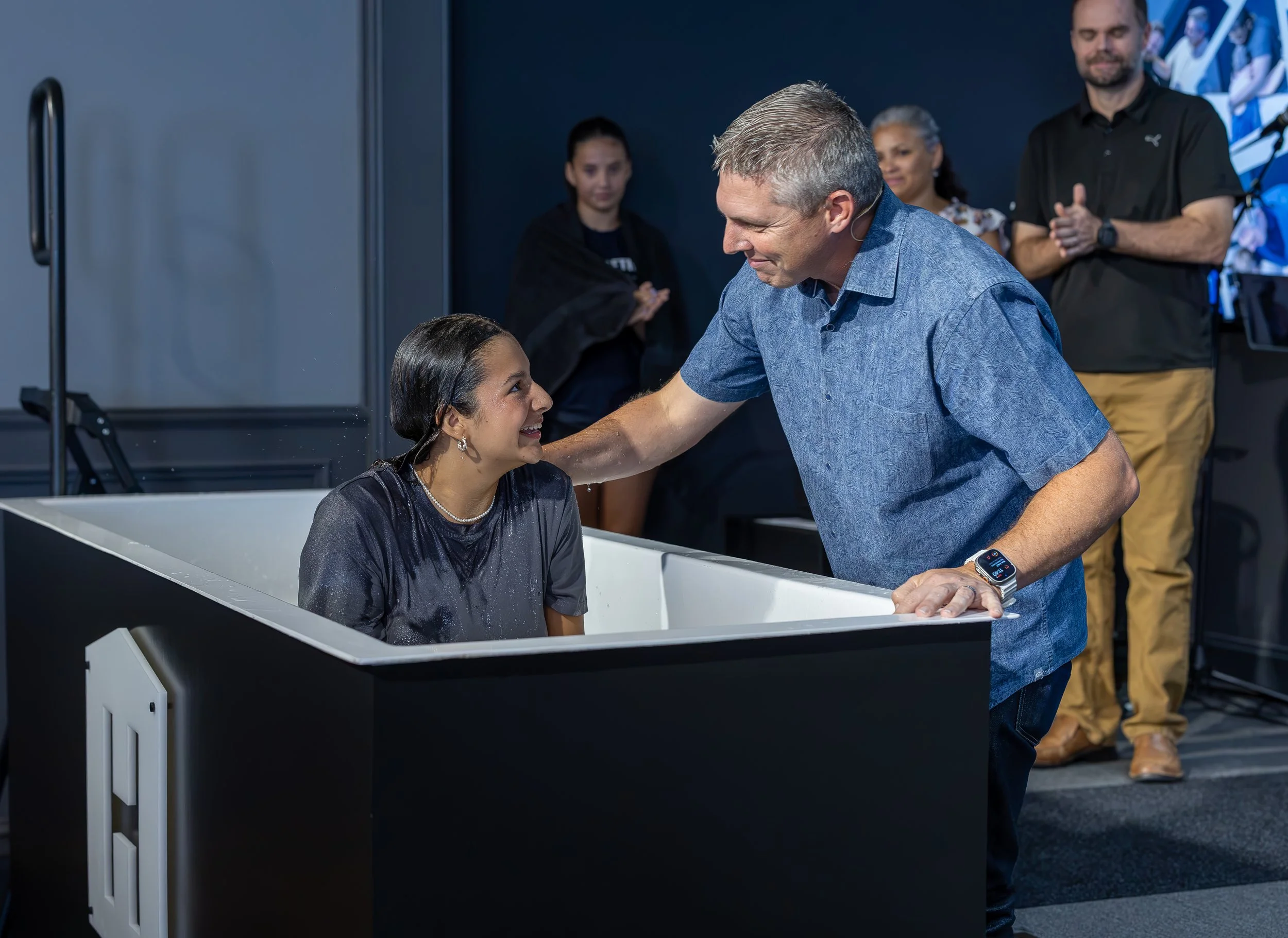 A man in a blue shirt is baptized in a small white baptismal pool by a smiling woman with earrings, while people watch in the background.