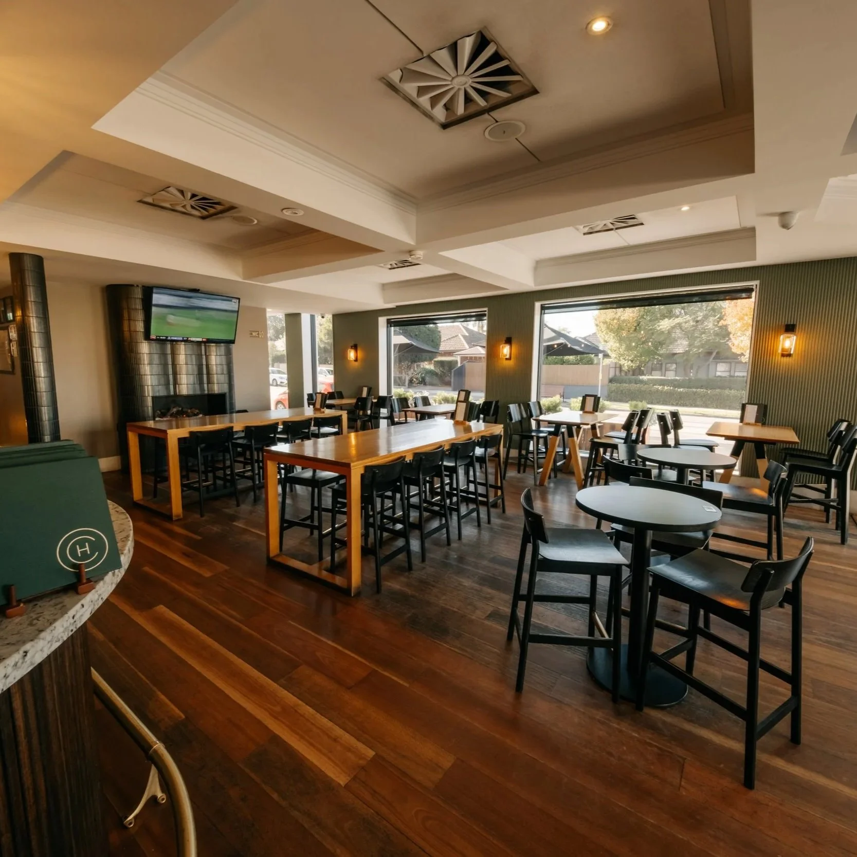 The Camden's Sports Bar with wooden tables and black chairs, large windows letting in natural light, wall-mounted TV, brick fireplace