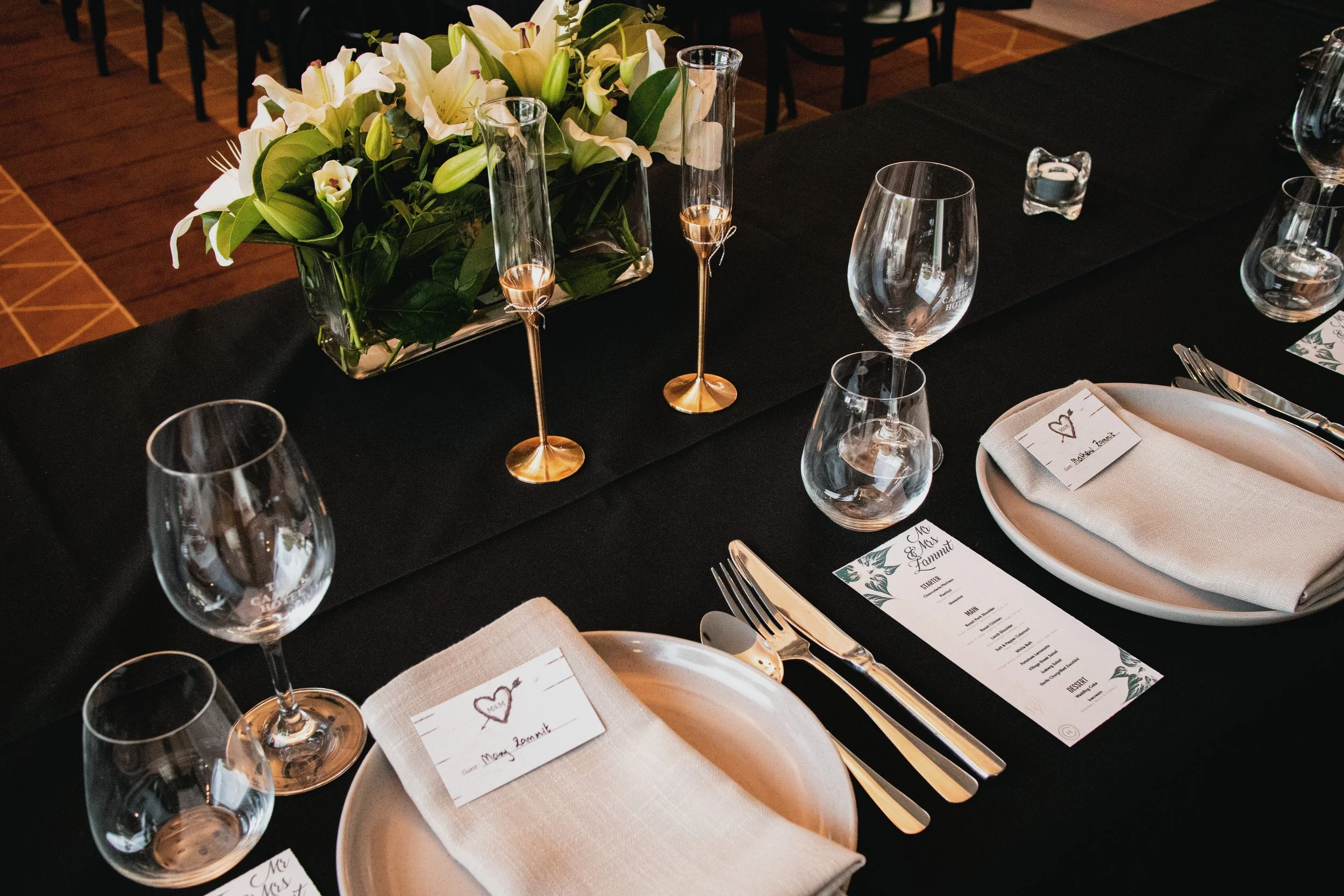 A table setting with a centerpiece of white lilies and greenery, surrounded by empty wine glasses, a menu card, a napkin with a handwritten note, and silverware, in a dimly lit room at The Camden