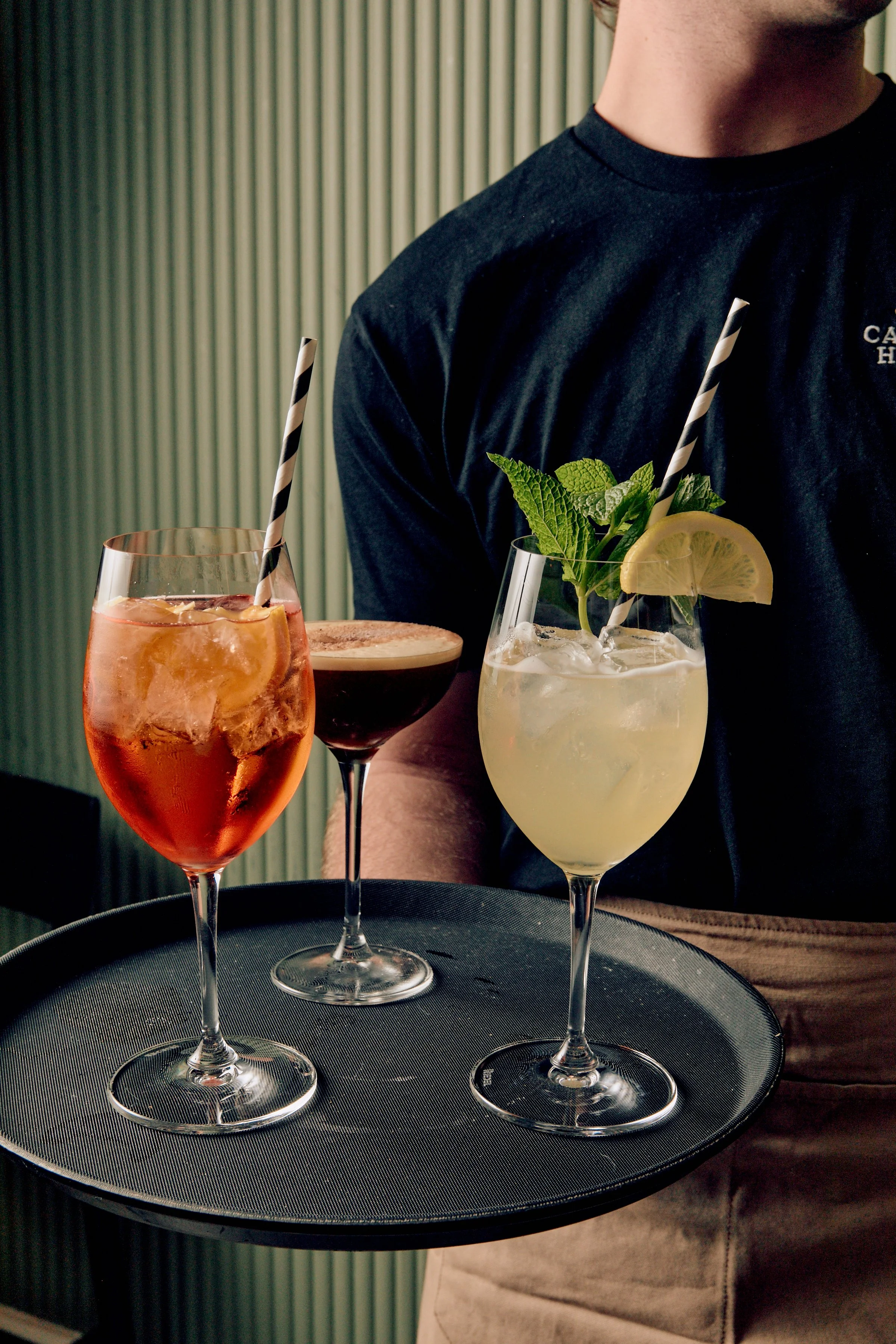 A person is holding a black tray with three colorful cocktails, each garnished differently, in a dimly lit setting with vertical blinds in the background.