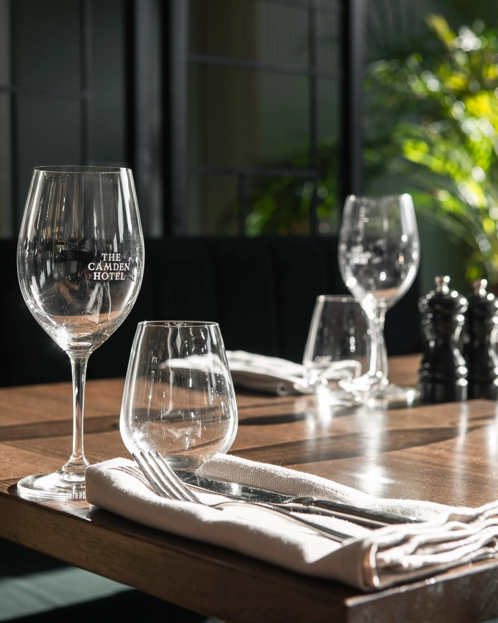 Empty wine glasses on a wooden table, with a napkin and cutlery, in a well-lit restaurant or cafe setting, with green foliage visible outside.