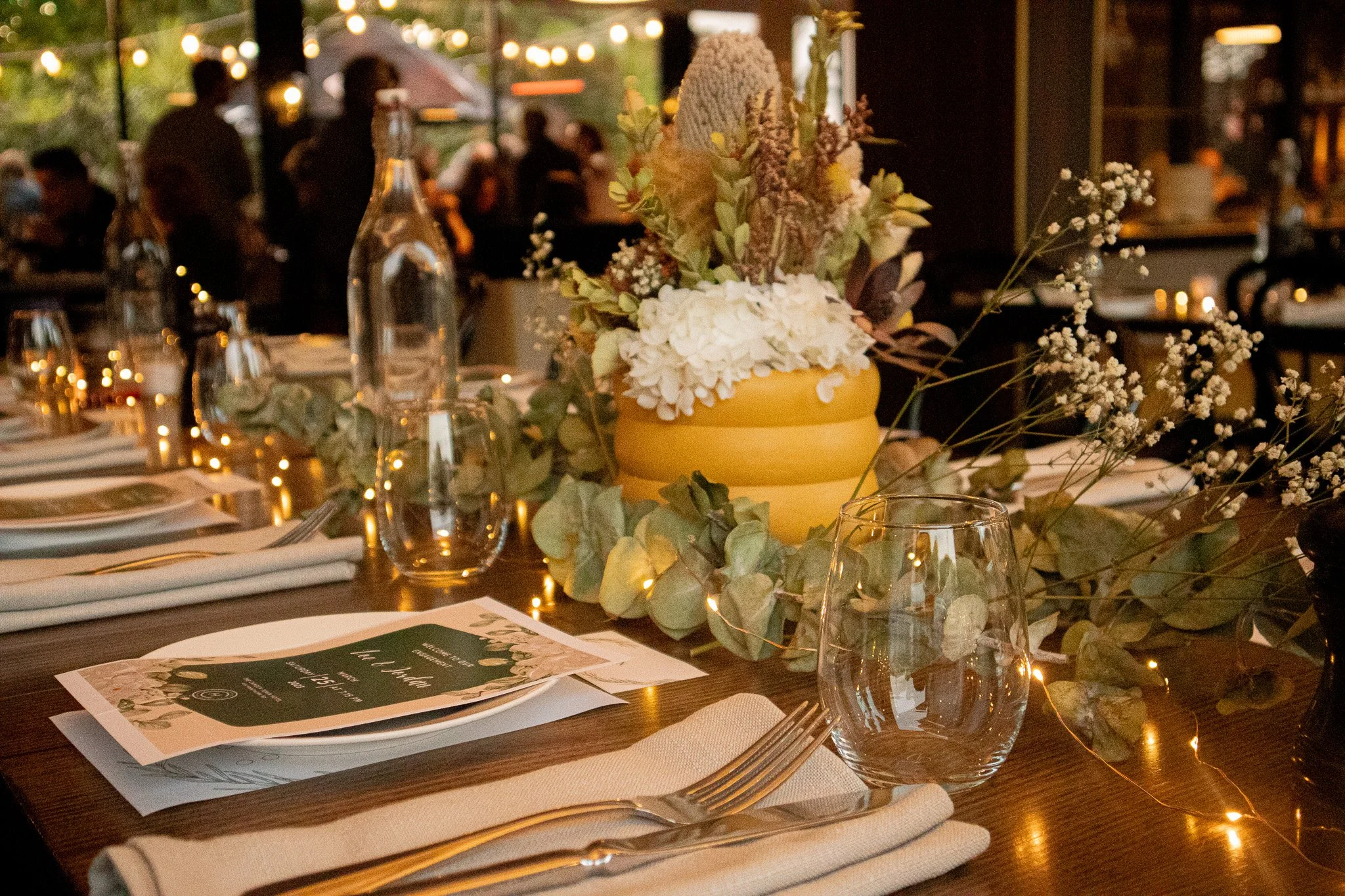 A table decorated with a bouquet of white and pink flowers in a yellow vase, surrounded by greenery, clear glass bottles, drinking glasses, place settings with white napkins, cutlery, and a menu, with soft warm lighting and blurred background of people at an indoor event.