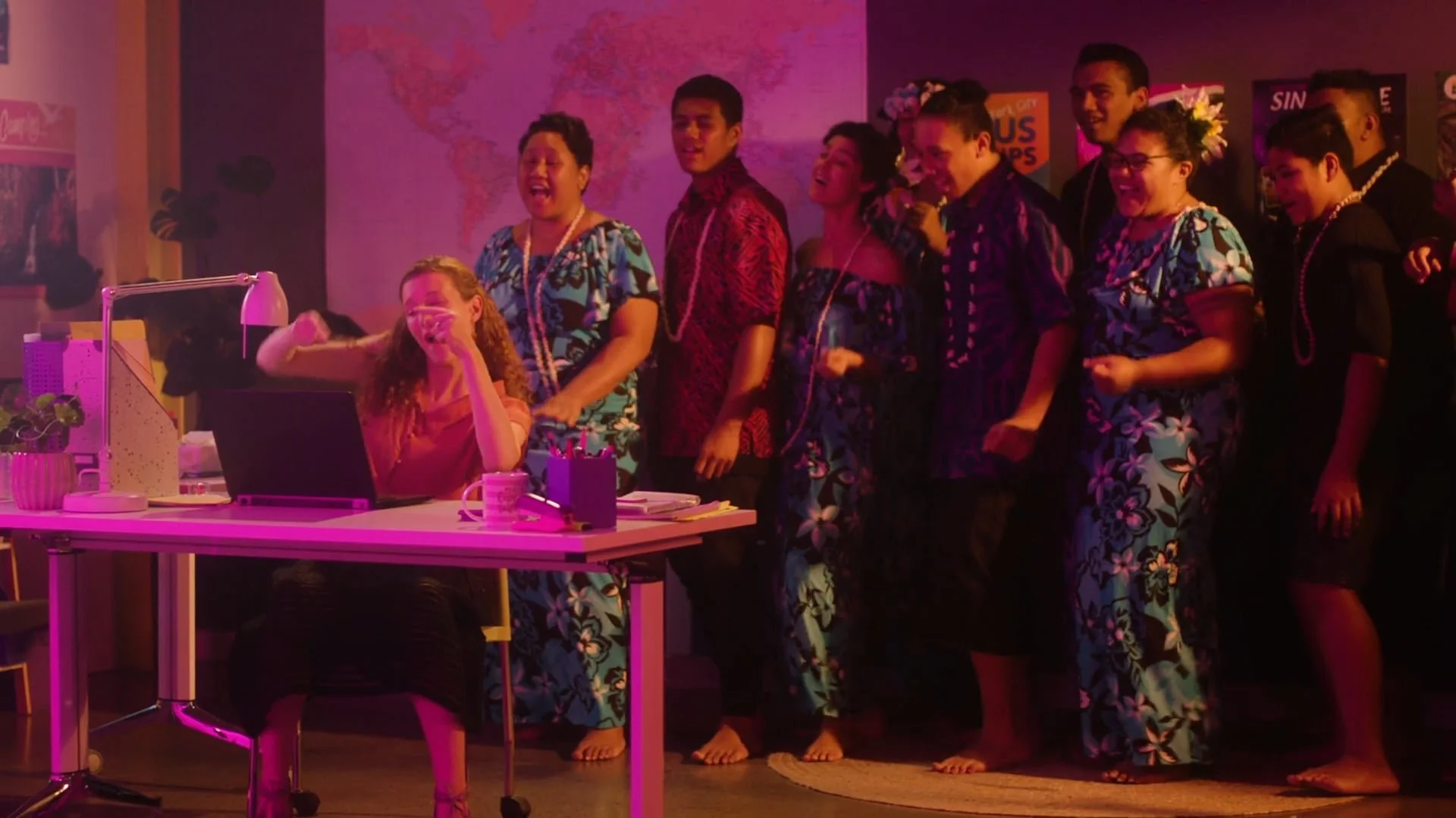A group of people in Hawaiian shirts and leis singing and dancing in an office setting with a woman at a desk using MYOB, colourful lighting, and a world map on the wall.