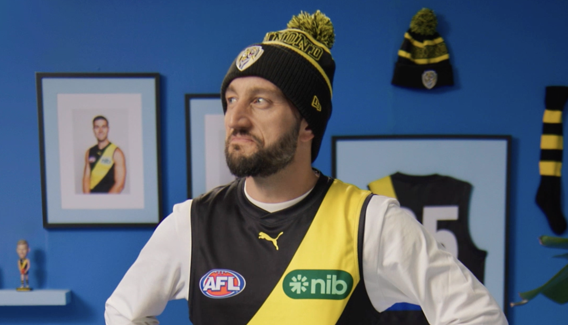 Troy the Tigers fan wearing the famous yellow and black sports jersey with AFL and nib logos, and a black beanie with yellow pom-pom and RFC logo, standing in a room with framed photos and sports memorabilia on blue walls.