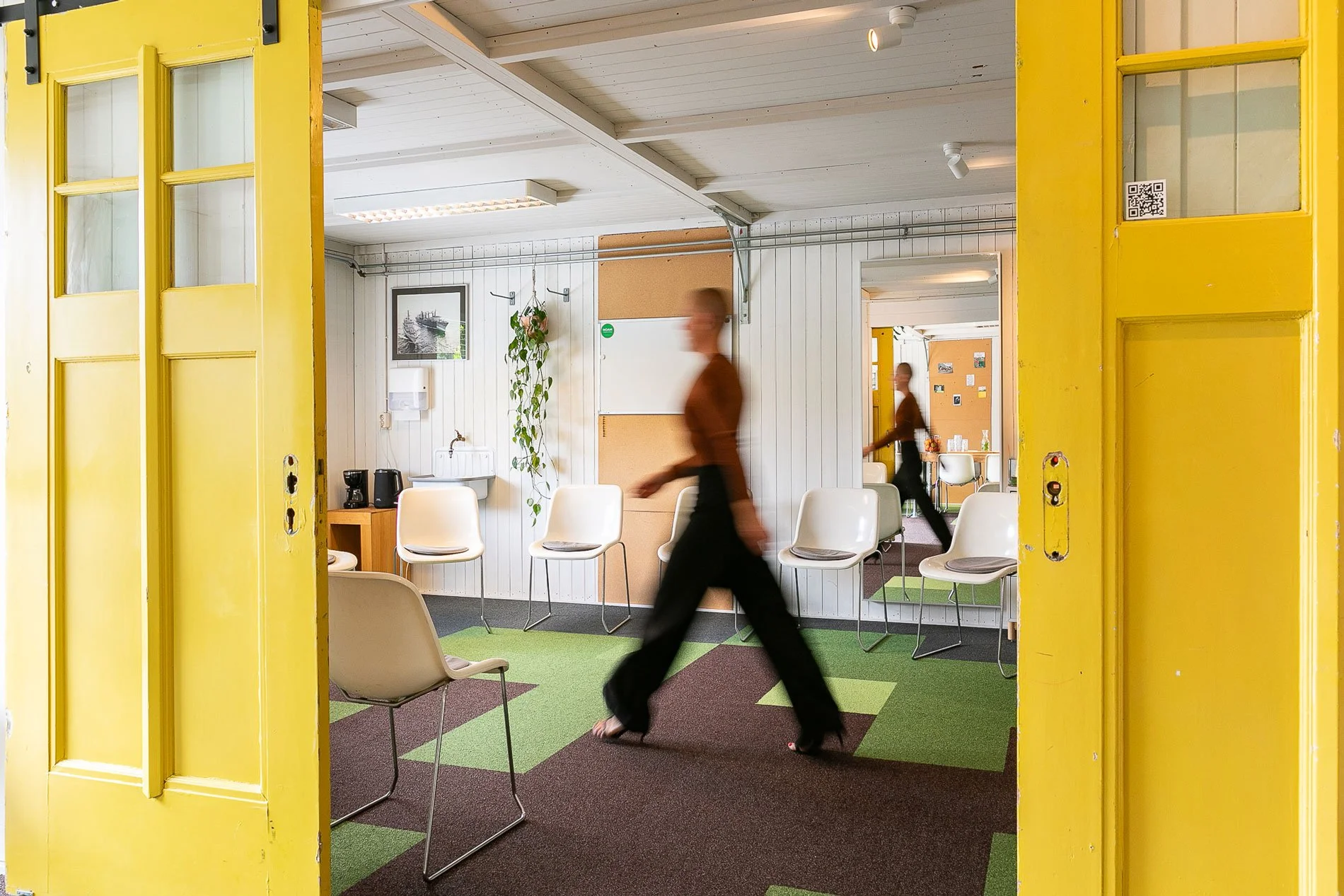 Room with white chairs, a small sink, coffee maker, hanging plant, framed photo, and a whiteboard. Two women walking through the space, viewed through an open yellow door.