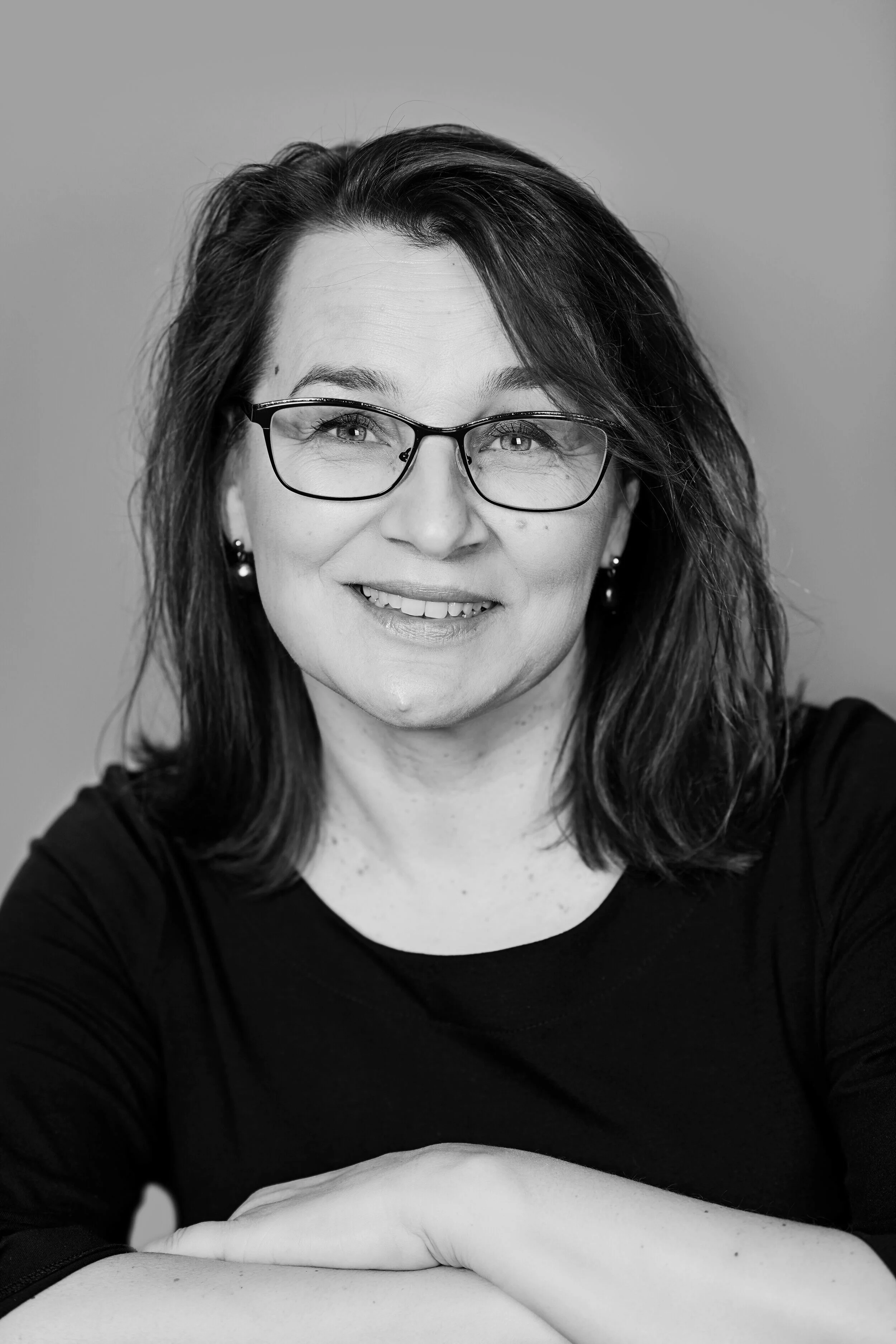 Musiikkiterapeutti Sari Hilpinen, hymyilee ja katsoo kameraan, hänellä silmälasit ja korvissa helmet. Hän istuu, käsivarsi lepää pöydällä.