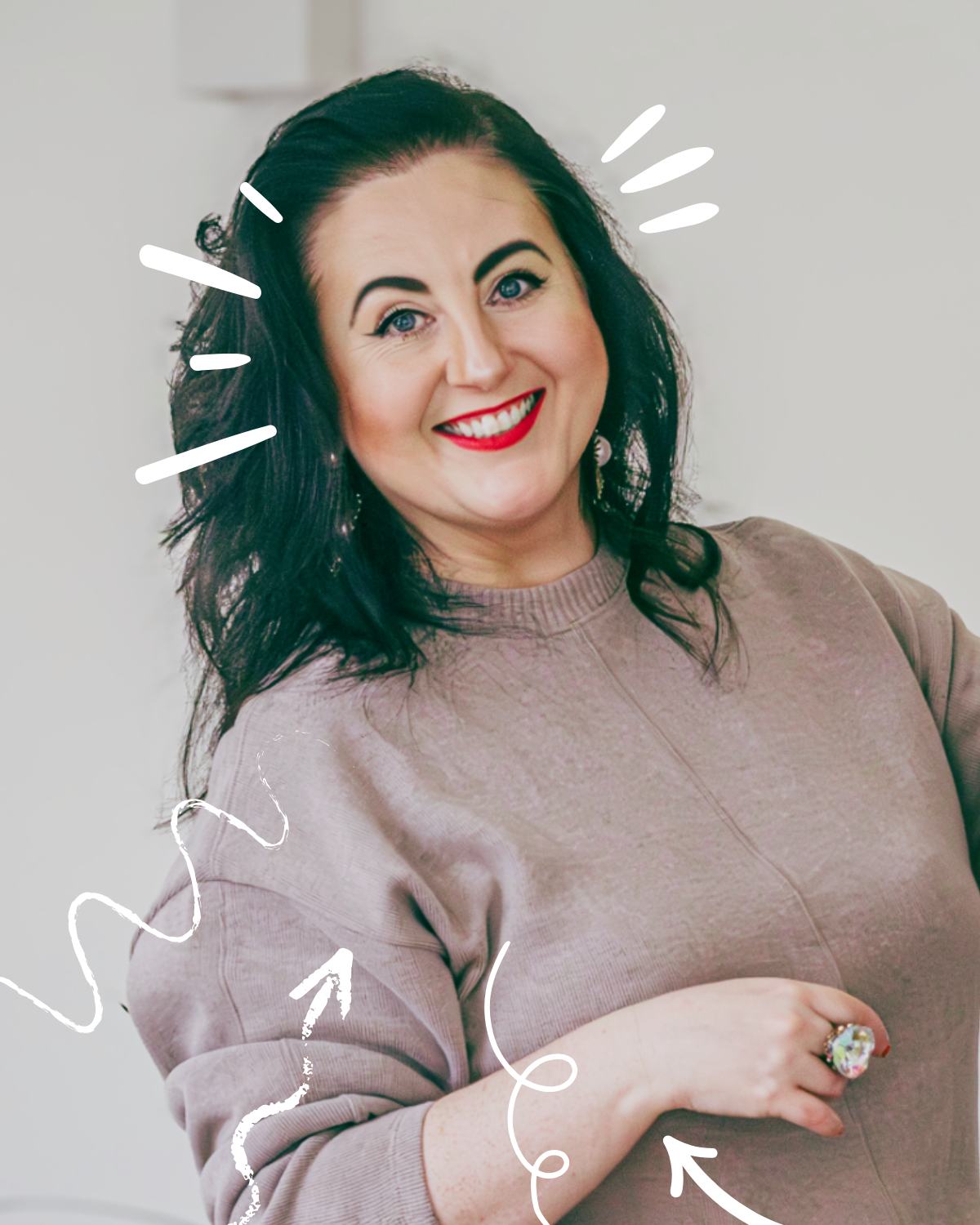 A woman smiling, wearing a beige sweatshirt, with dark hair and bright lipstick, with white decorative lines and arrows drawn around her face and arm.