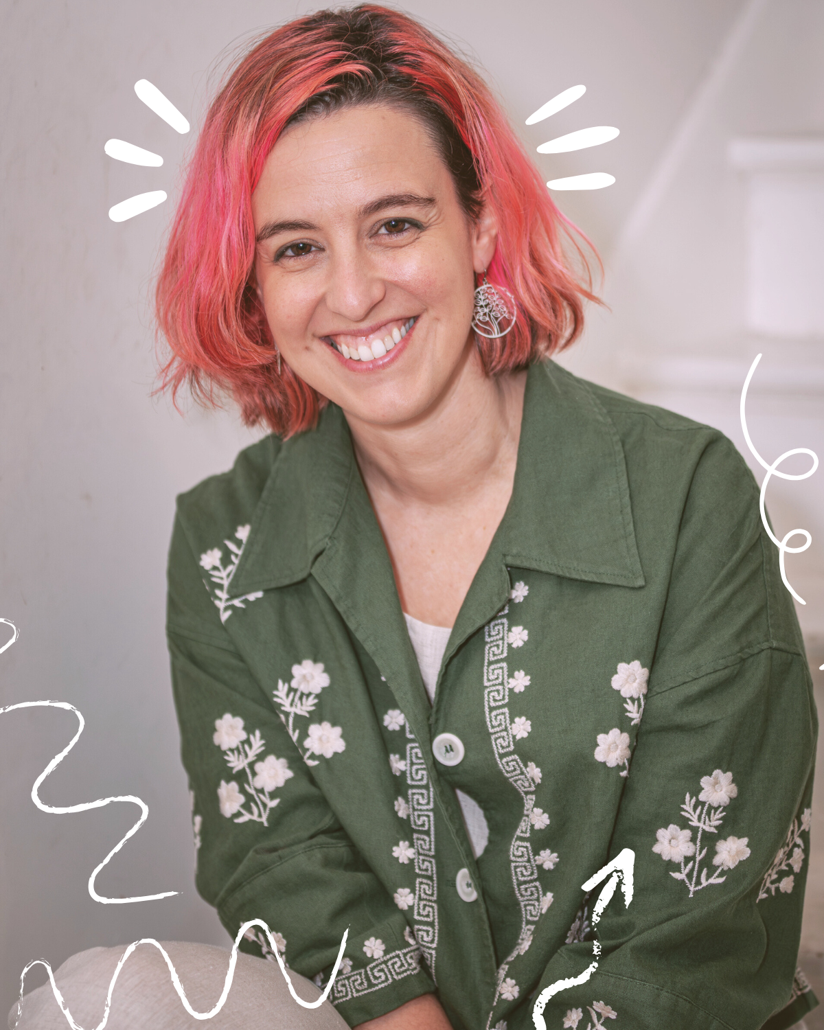 A woman with pink hair and earrings smiling, wearing a green jacket with white floral patterns, sitting indoors.