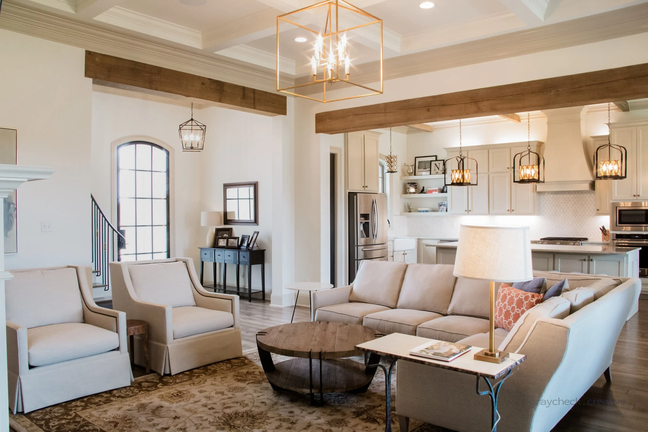Bright living room with beige sofa, two matching armchairs, wooden coffee table, cream walls, patterned rug, white kitchen with pendant lights in background.