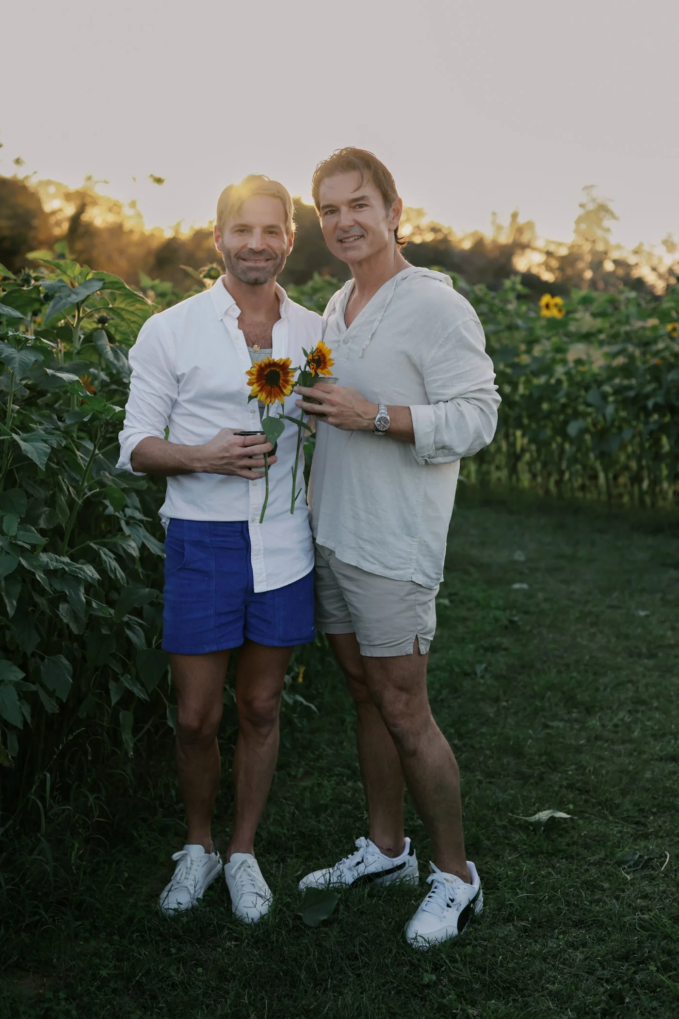 Two men standing in a sunflower field at sunset, holding sunflowers and drinks, dressed casually in shorts and shirts.