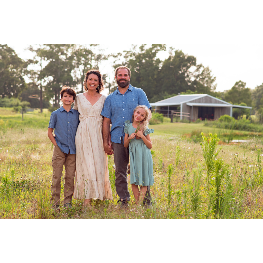 A family of four standing outdoors in a grassy field, smiling at the camera, with a barn and trees in the background.